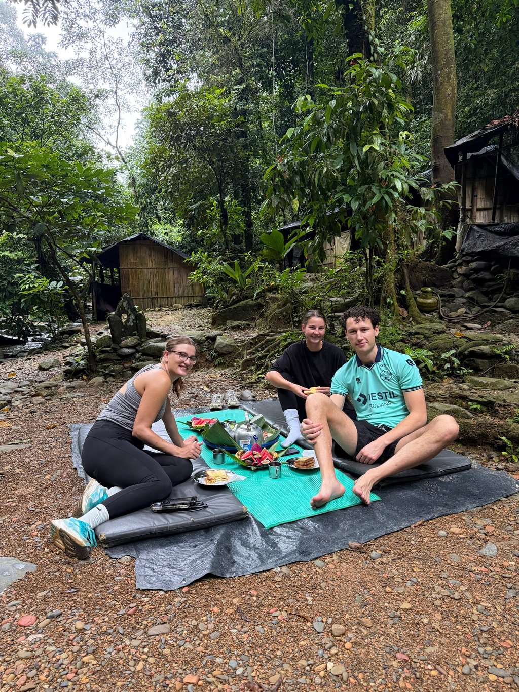 Young travelers enjoying a jungle picnic on mats near rustic wooden cabins in a tropical forest.