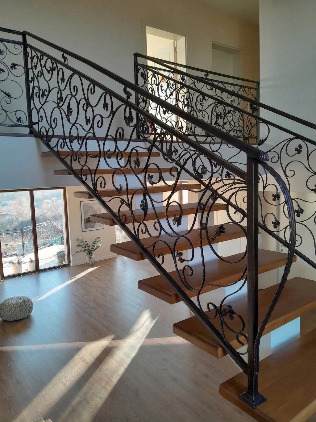 Modern floating wooden staircase featuring ornate wrought iron scrollwork railings in a bright, sunlit home.