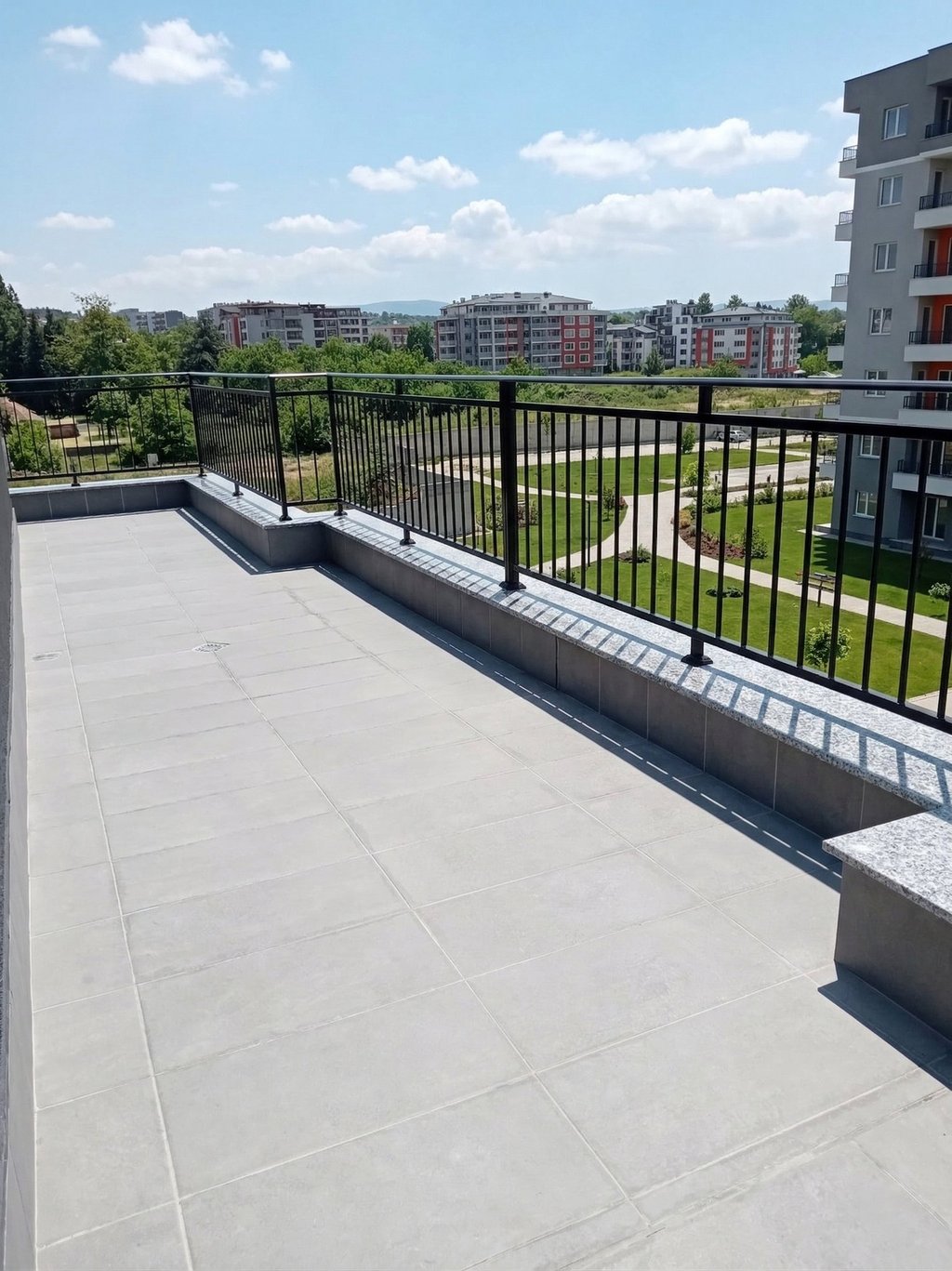 Modern apartment balcony with grey tile flooring and black metal railing overlooking a landscaped park.