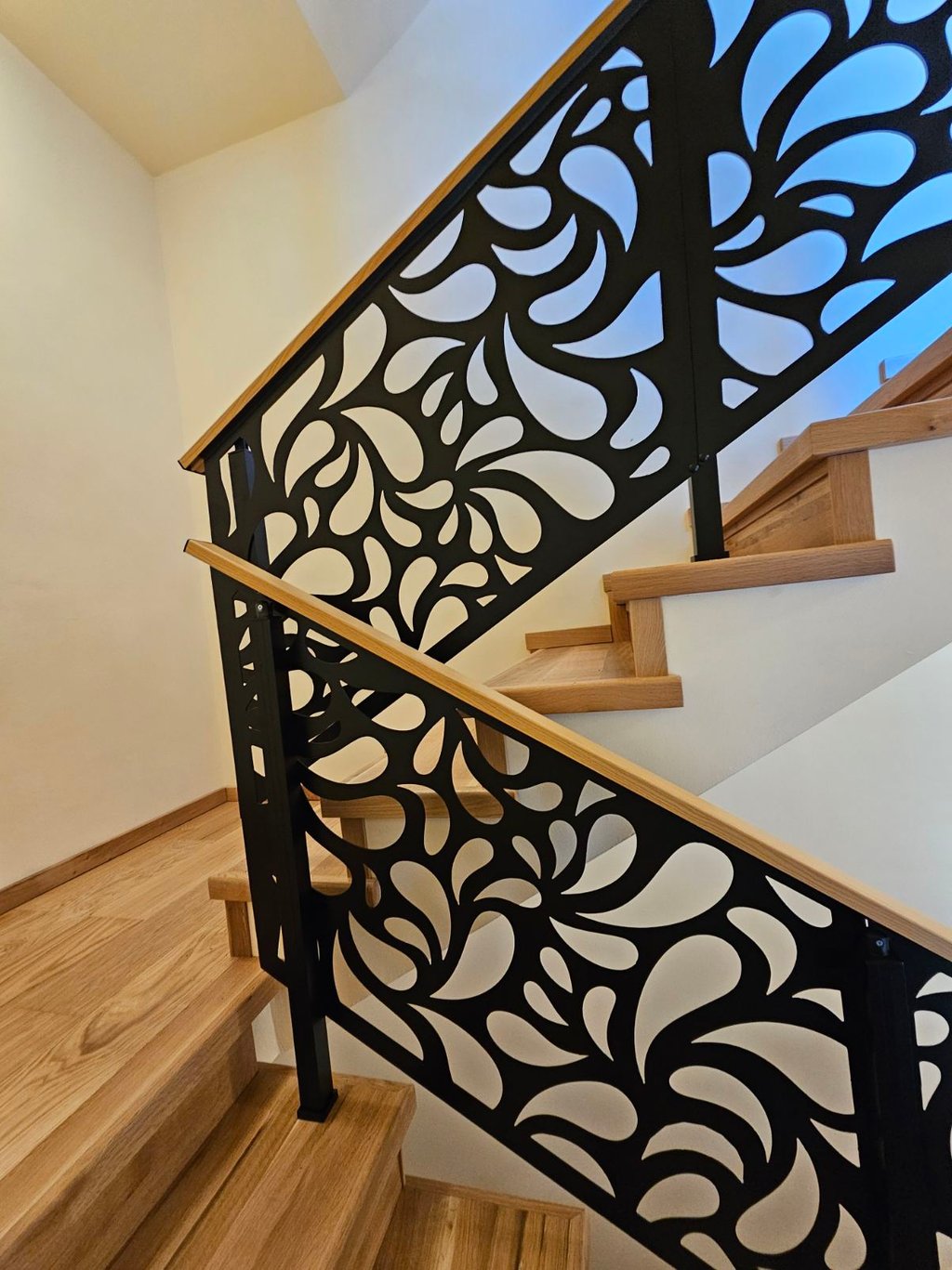 Modern wooden staircase featuring decorative black laser-cut metal railings with a floral swirl pattern.