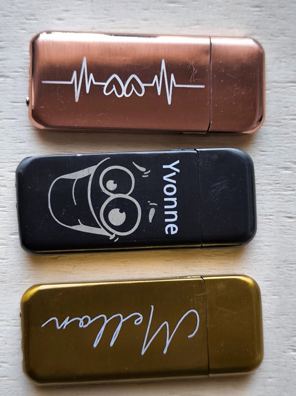 Three personalized rechargeable lighters in rose gold, black, and gold finishes with custom laser engravings.