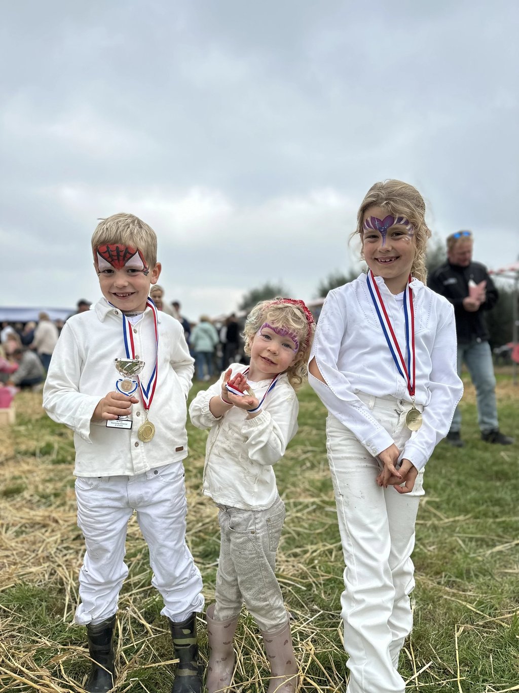 Drie trotse kinderen met medailles en schminkcreaties op een evenementenveld.
