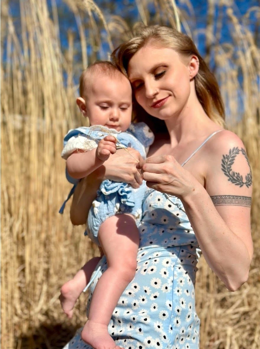 A mother and baby girl both wearing matching blue dresses in a hay field on a sunny day, symboling maternal love.