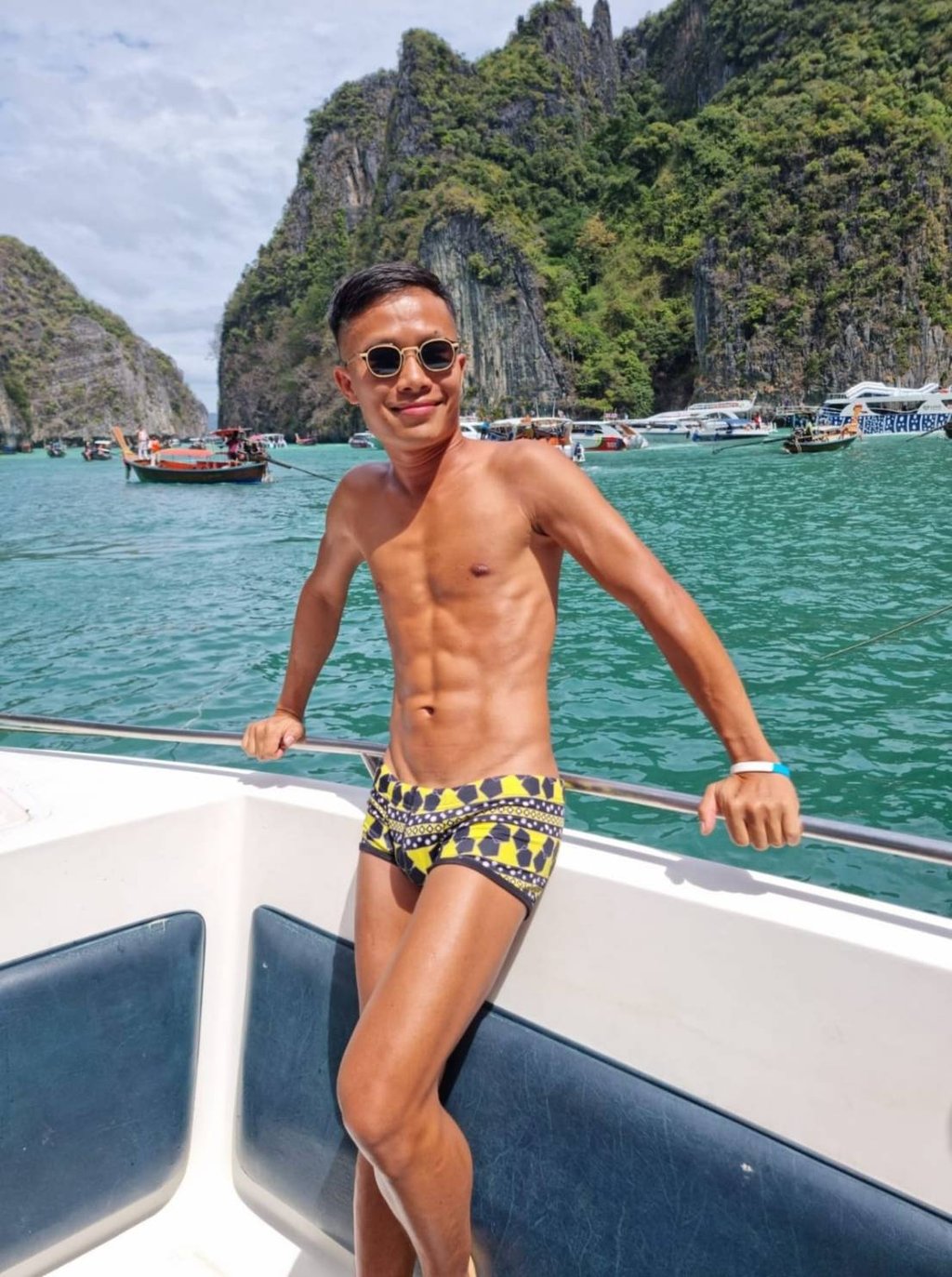 A male outcall masseur in Pattaya standing on a boat wearing swim wear with the sea and an island in the background