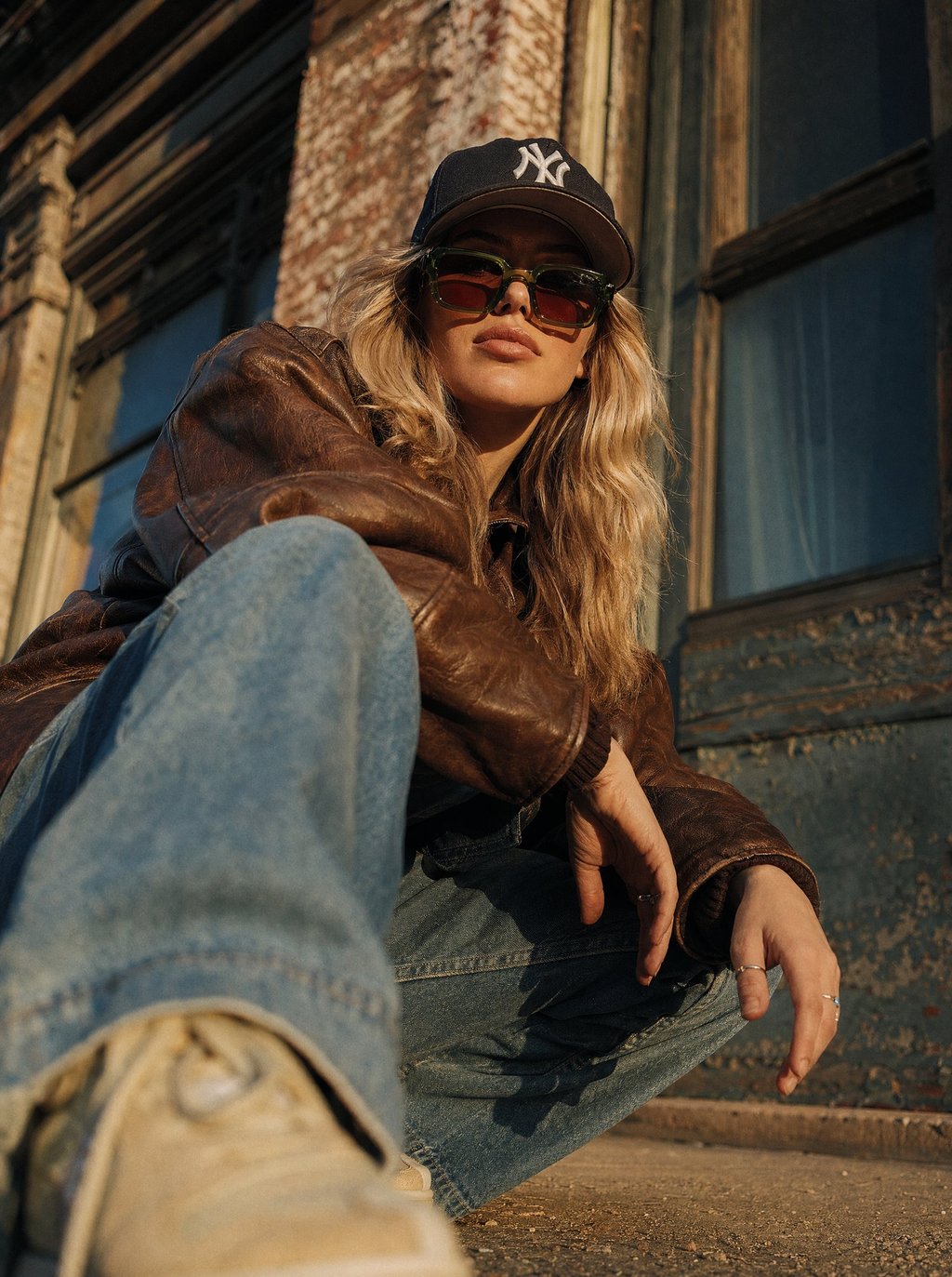 A blonde woman in a brown leather jacket, sunglasses, and baseball cap posing in urban street style.