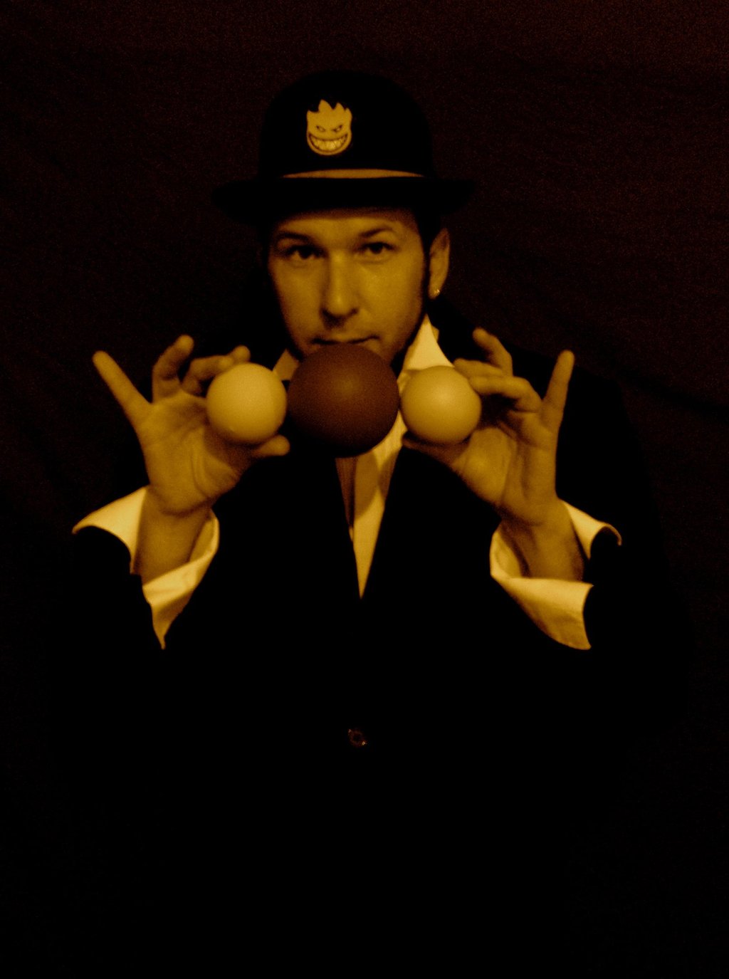 a man in a suit and hat with two hands up to the camera holding juggling balls