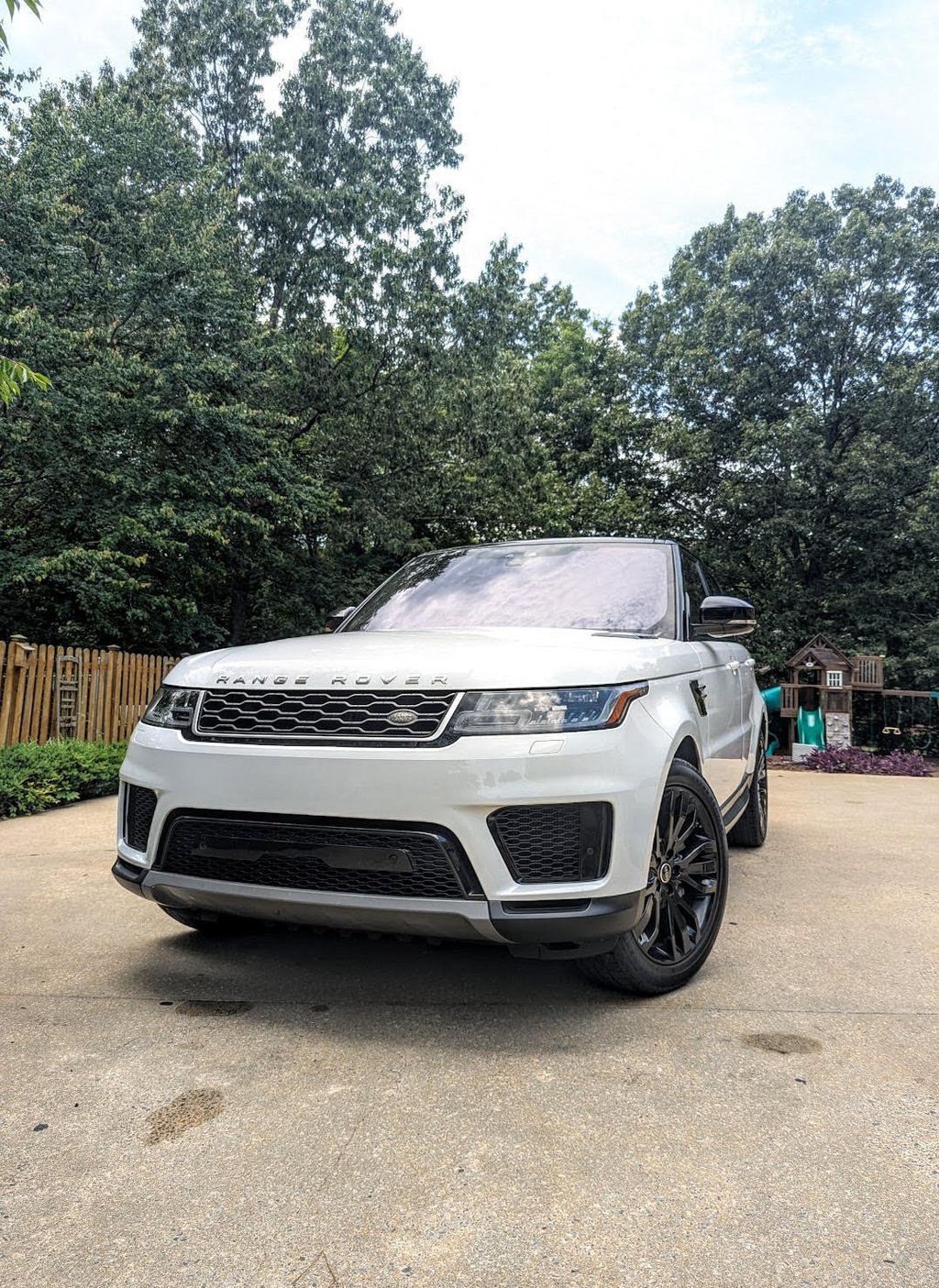 White Land Rover Range Rover Sport with black rims parked on a driveway near green trees.