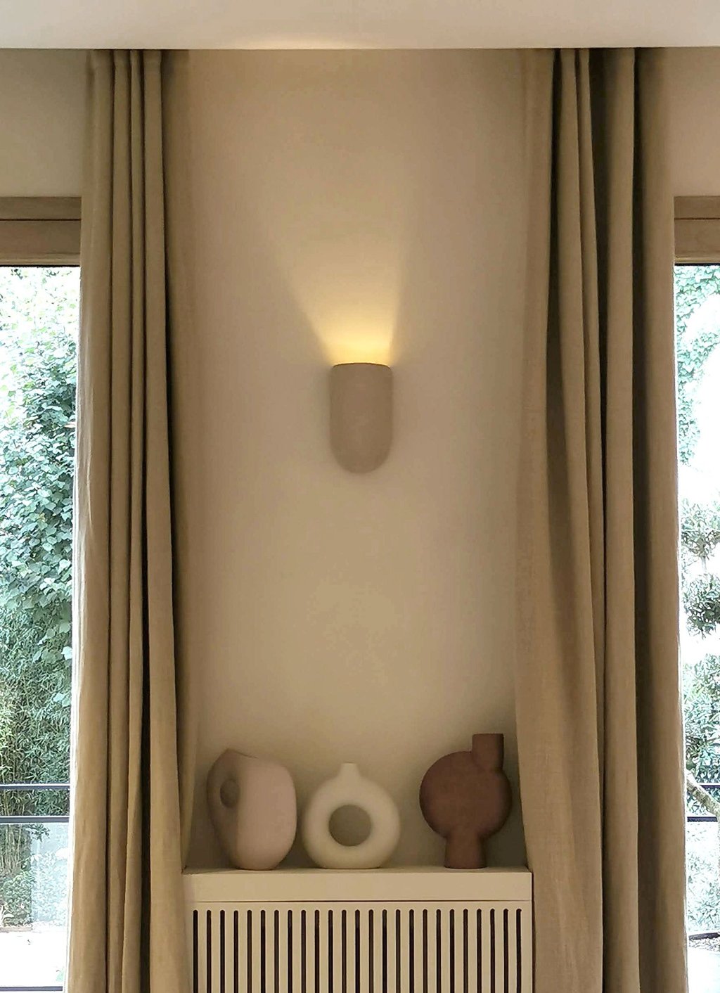 Modern minimalist wall sconce lighting over ceramic vases and neutral linen curtains.