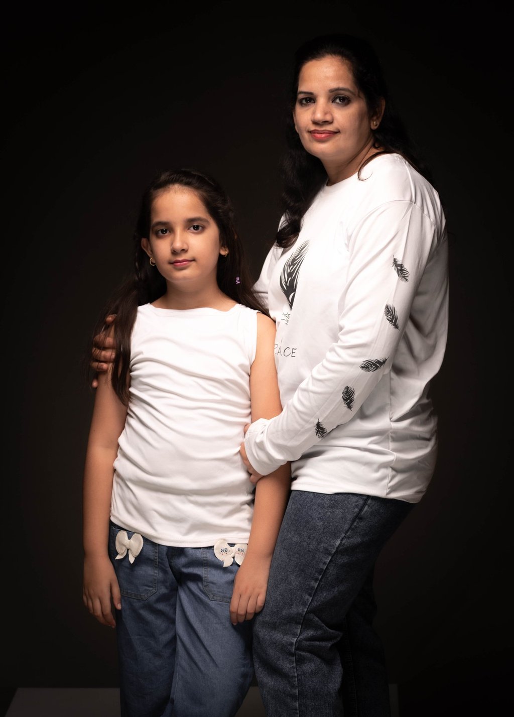 A portrait of a mother and daughter posing together in white shirts and denim jeans against a dark background.