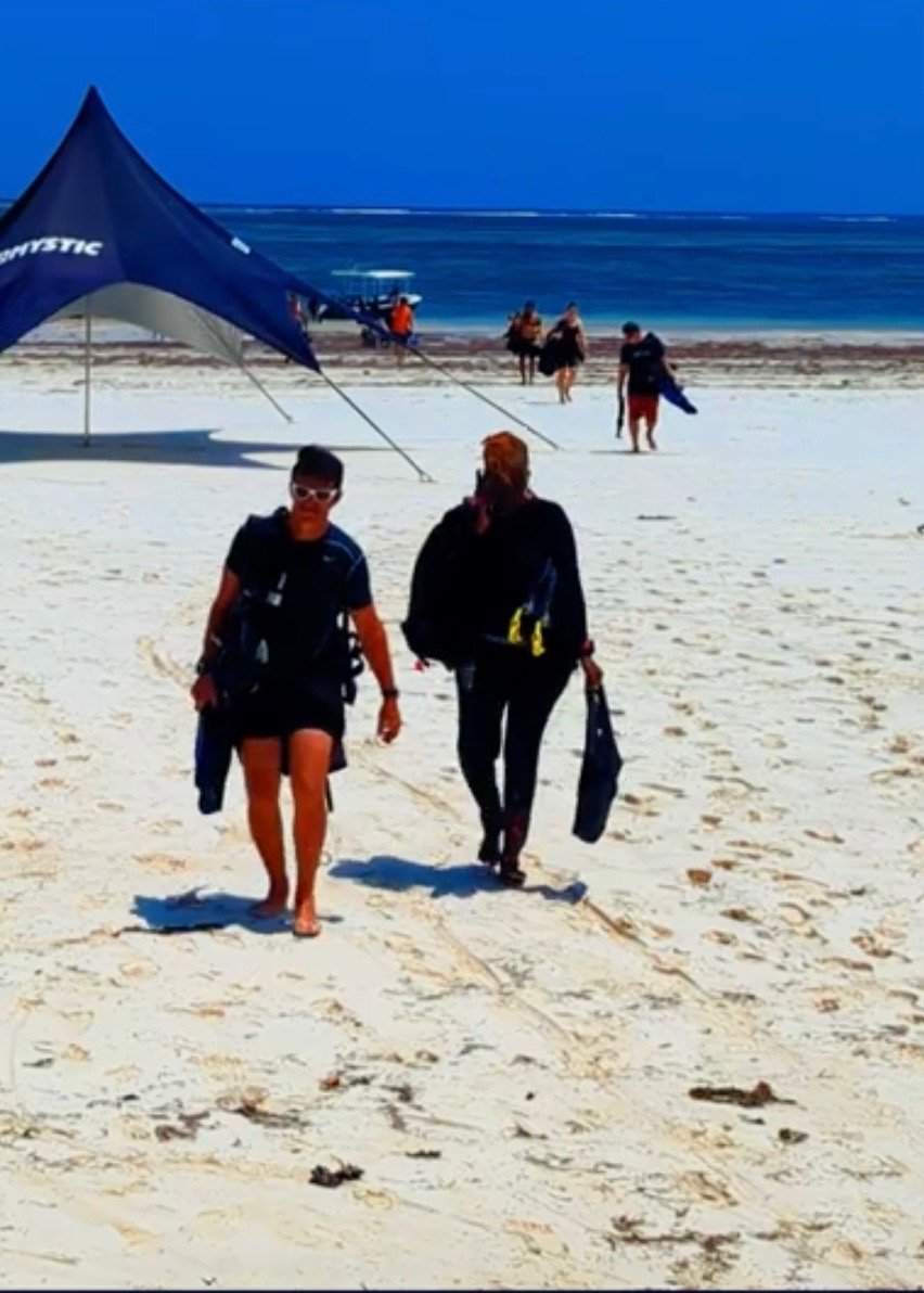 Scuba divers walking on a white sand tropical beach carrying diving gear near a blue ocean.