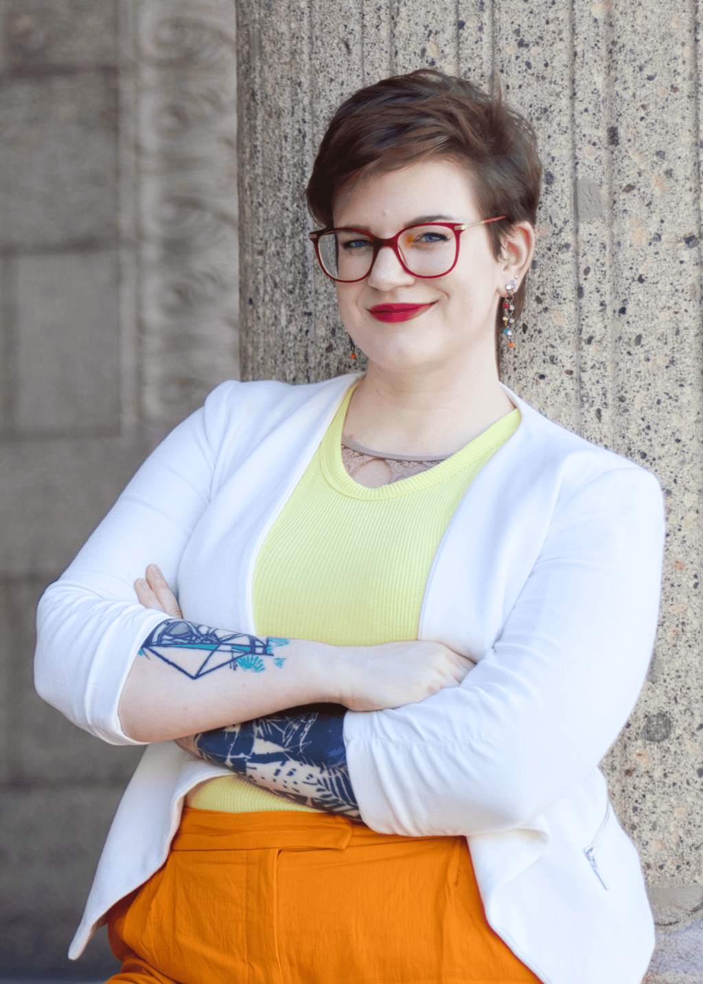 Swana Schuchmann mit Tattoos auf ihren Armen, roter Brille, gelben Top und weißem Blazer, lächelnd