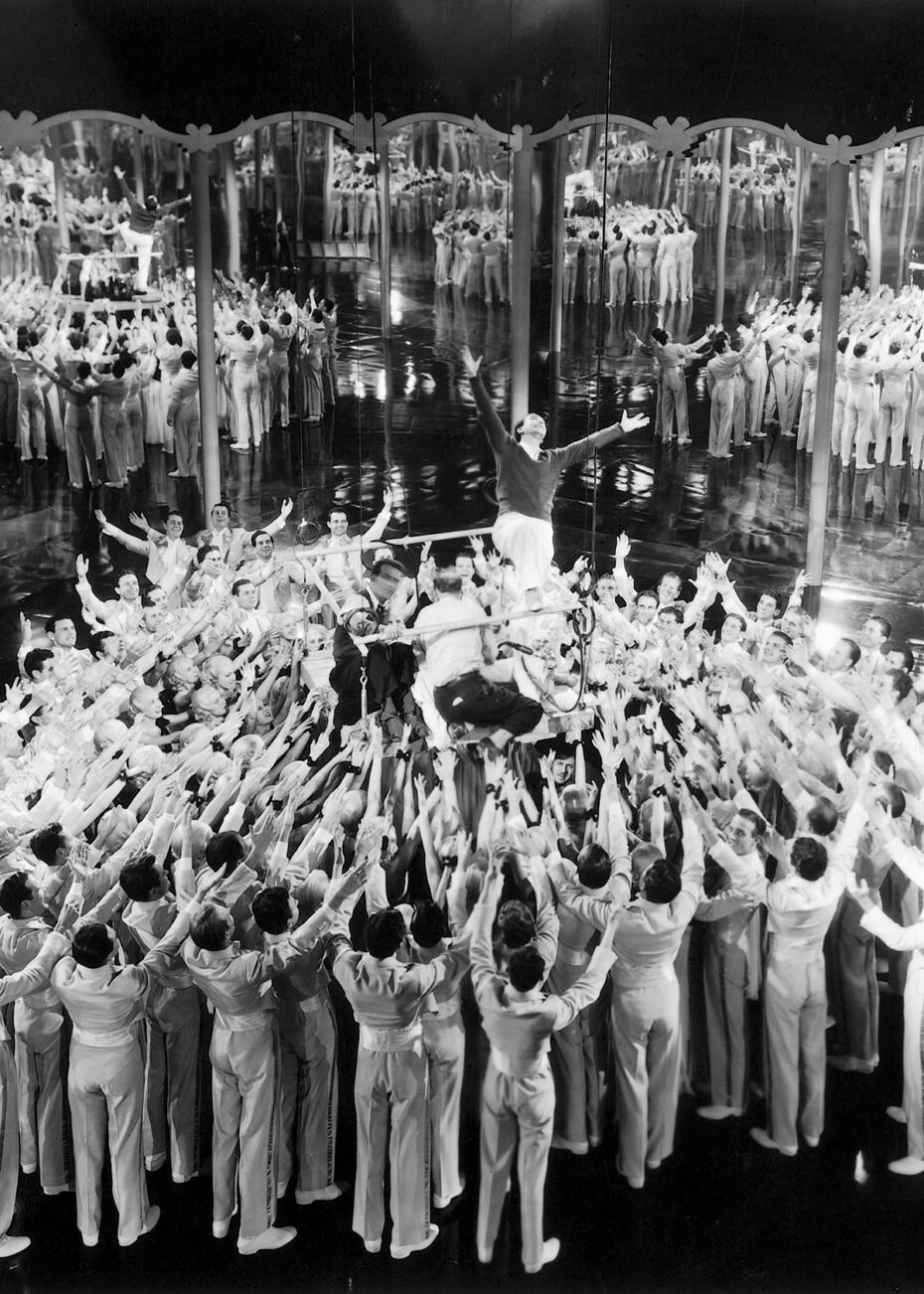 Busby Berkeley demonstrating arm movements to dancers on a mirrored stage platform.
