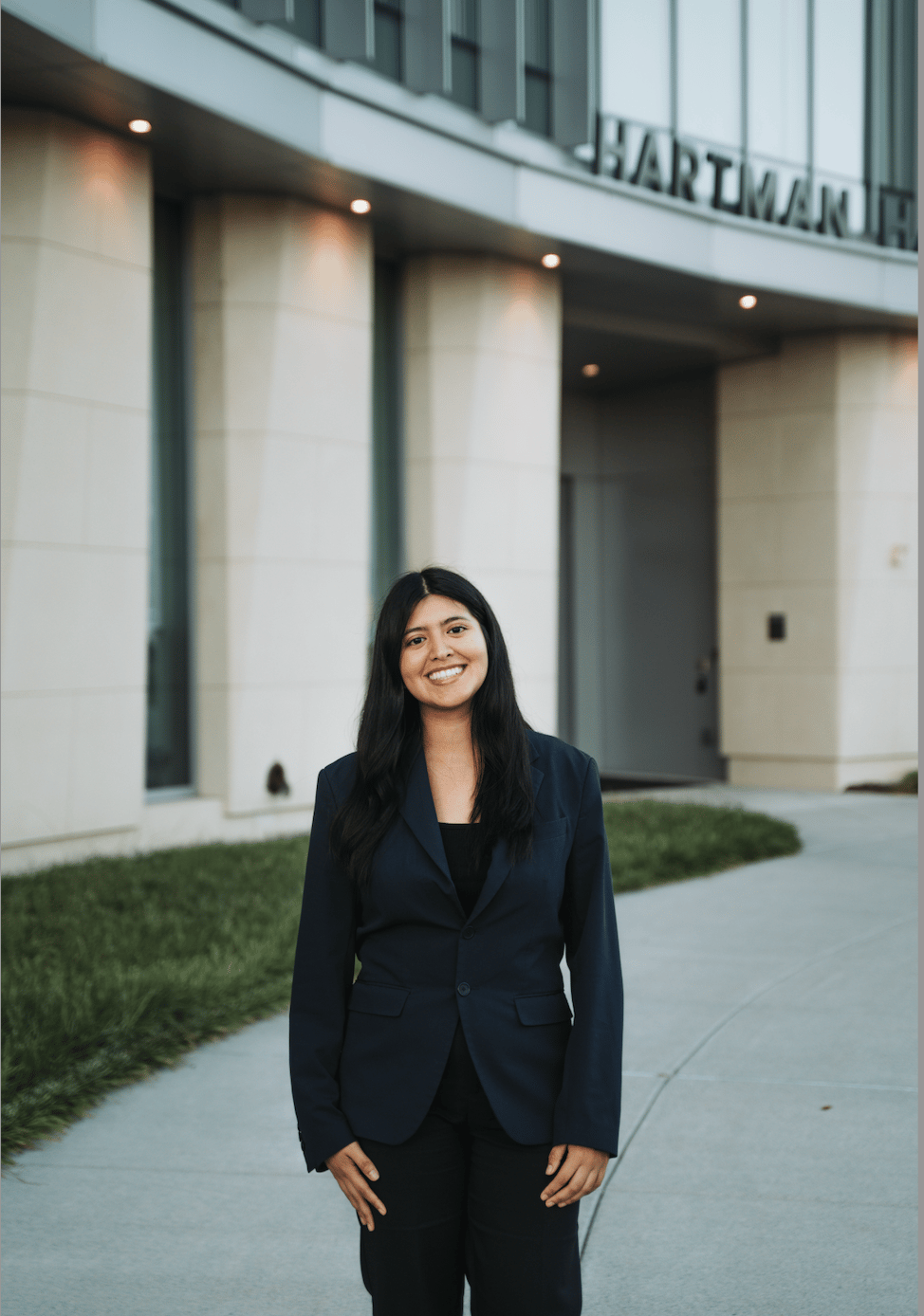 Professional woman in a navy blazer smiling in front of the Hartman Hall building on campus.