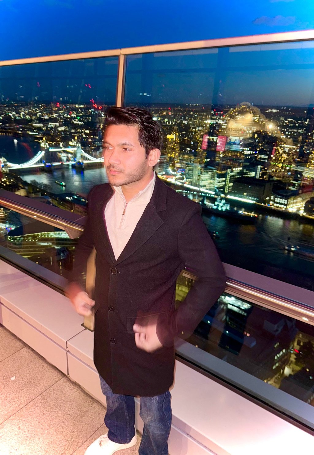 A man in a black coat standing on a London rooftop balcony at night with Tower Bridge in the background.