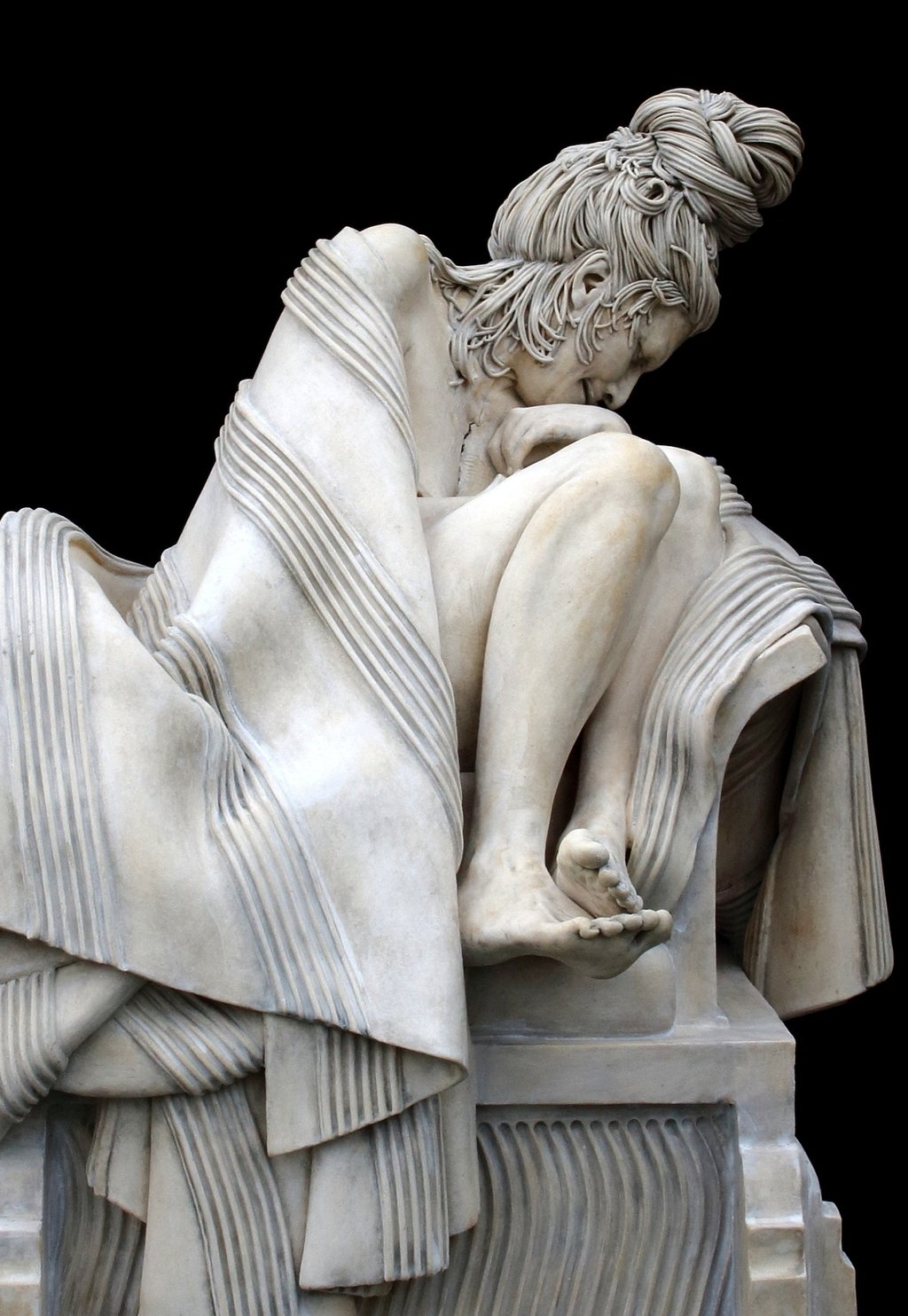Closeup front of ceramic sculpture of grieving woman wrapped in blanket on large classical plinth.