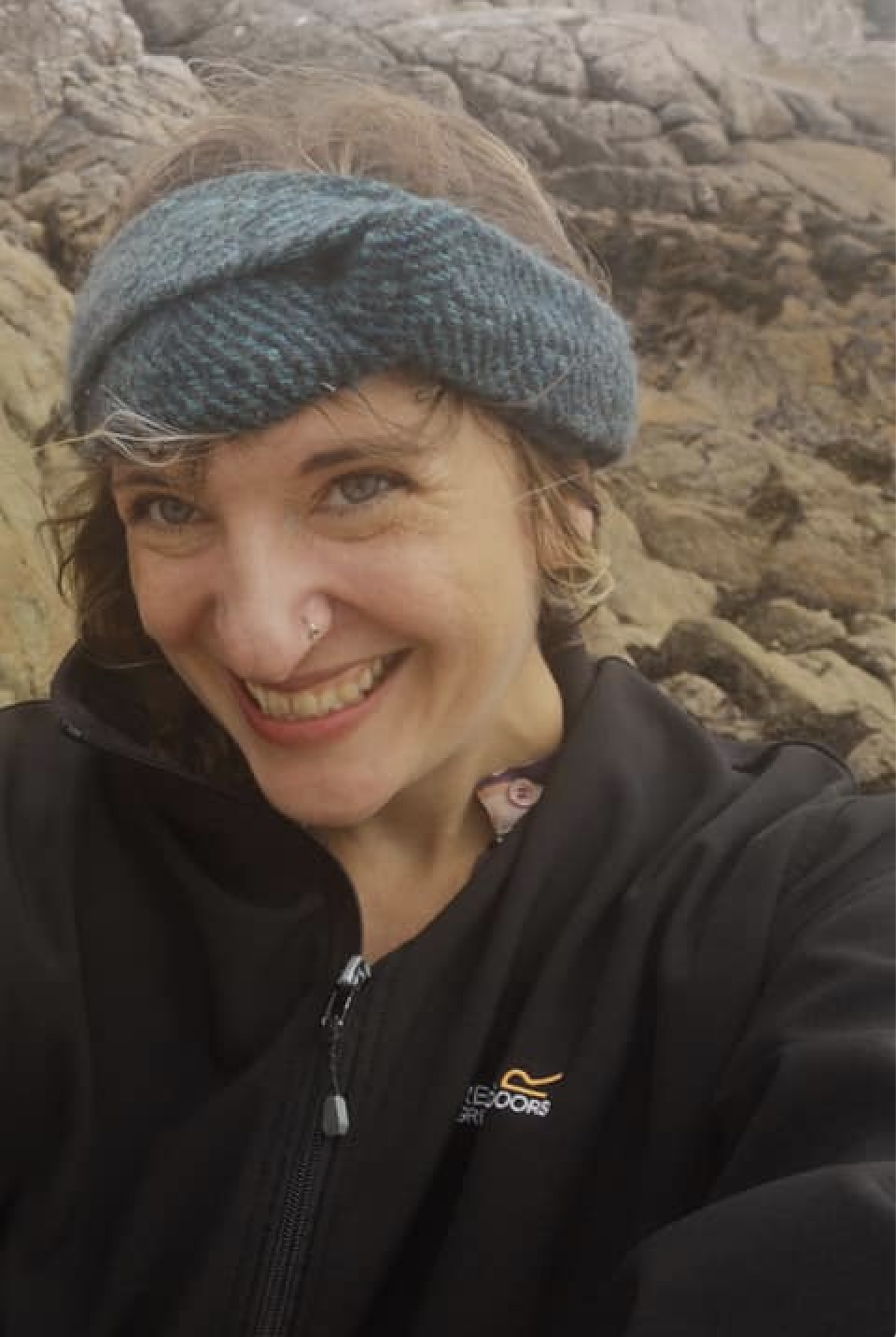 Smiling woman wearing a blue knit headband and black jacket standing on a rocky shoreline.