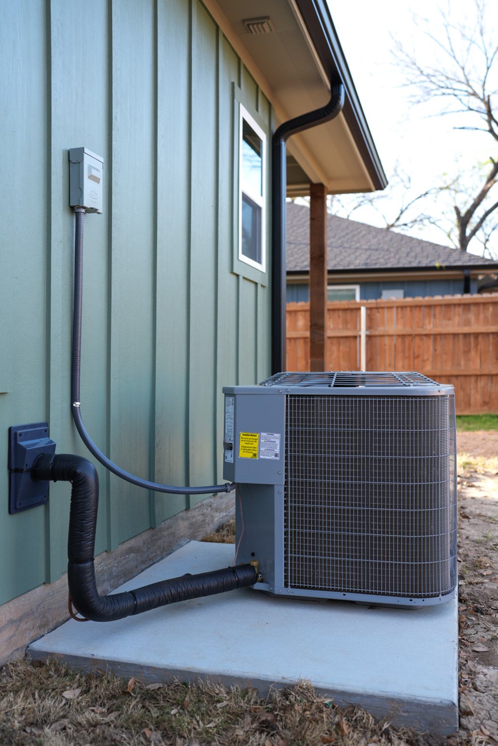 Exterior residential HVAC condenser unit installed on a concrete pad next to a green house wall.