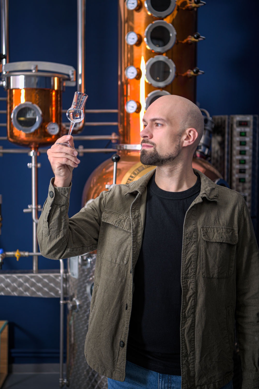 mastro distillatore che osserva del liquido in un bicchiere da degustazione davanti a un alambicco
