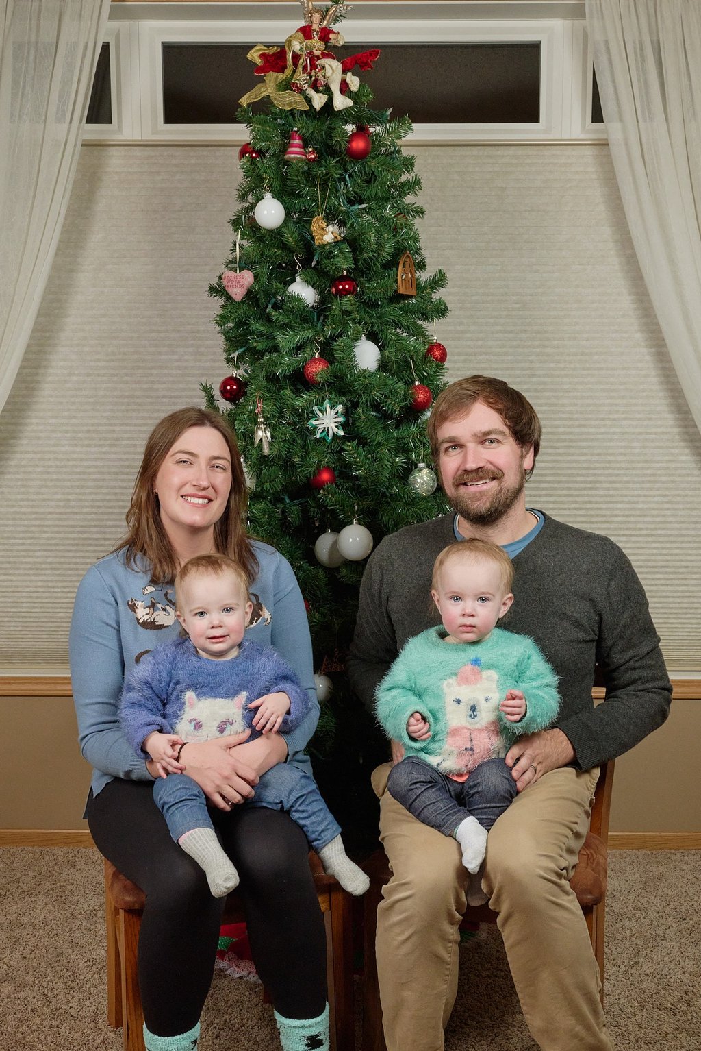 A happy family of four with twin babies sitting in front of a decorated Christmas tree.