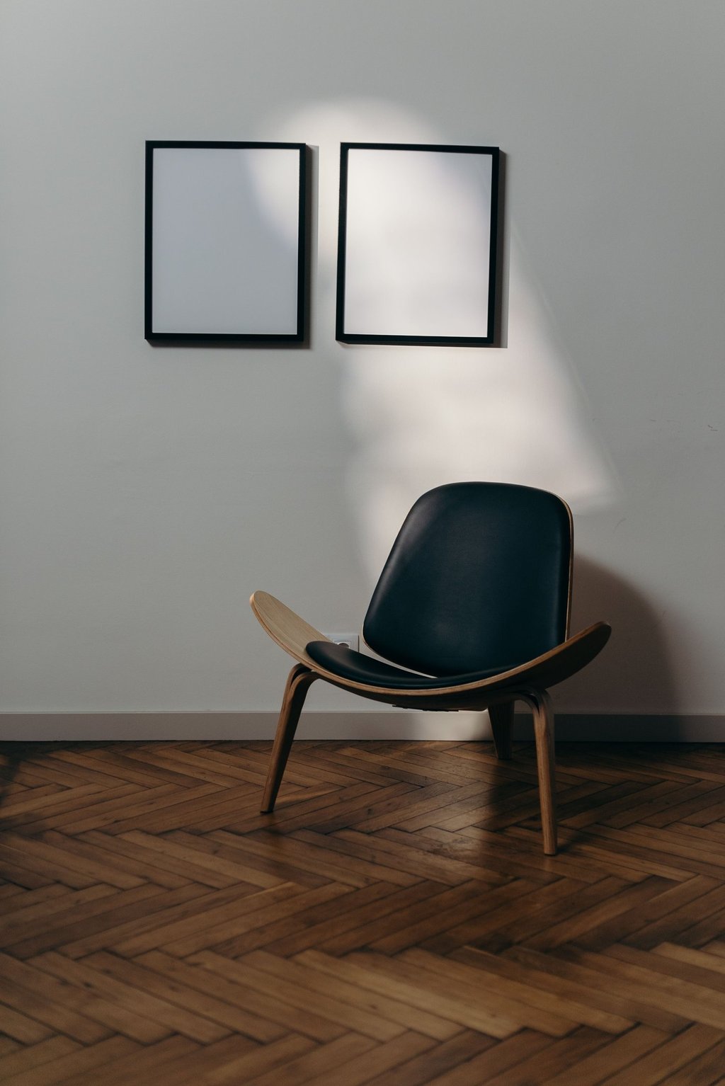 Modern black leather shell chair on herringbone wood floors with blank wall art frames in a minimalist room.