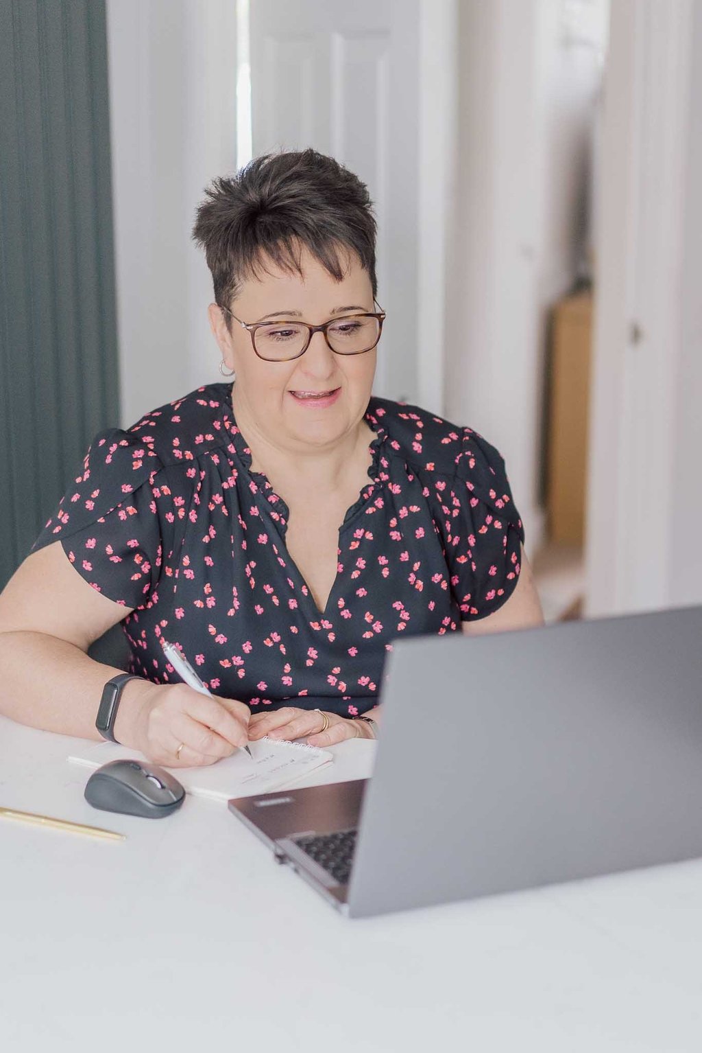 Professional woman with glasses working on a laptop and taking notes in a home office.