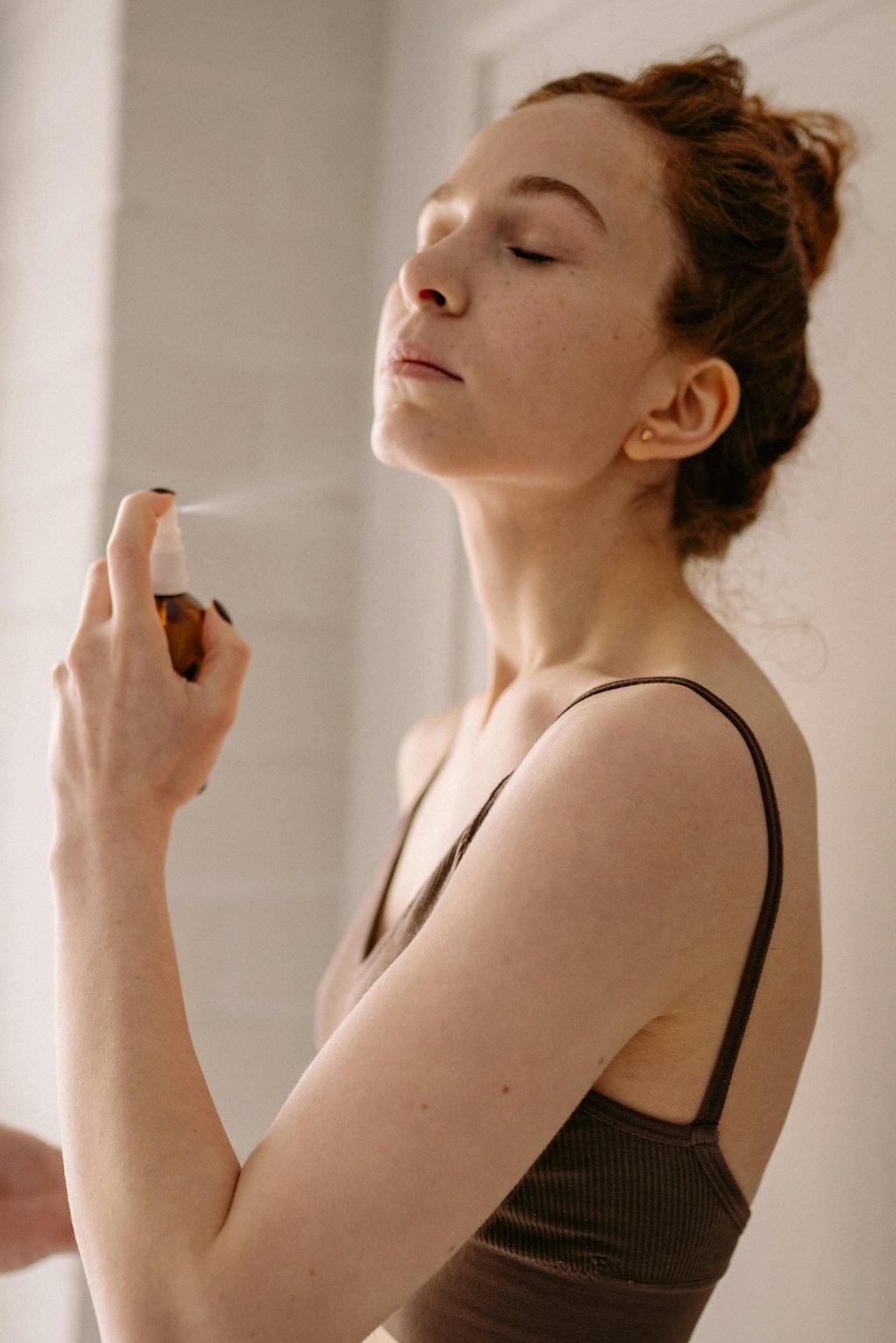 a woman in a brown tank top spraying a facial mist on her face
