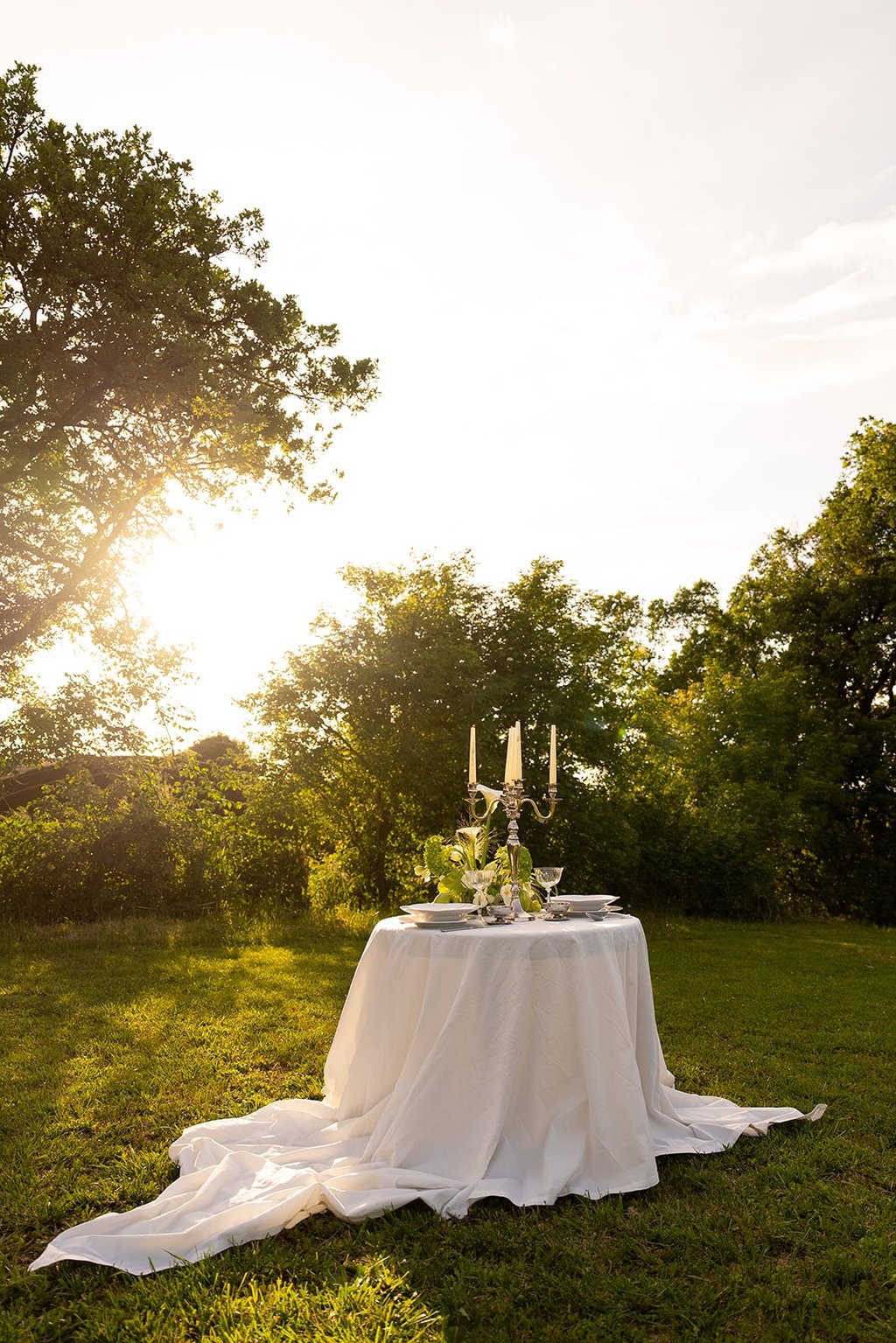 Elegant outdoor dinner table set with a white tablecloth and candelabra in a sunlit garden.