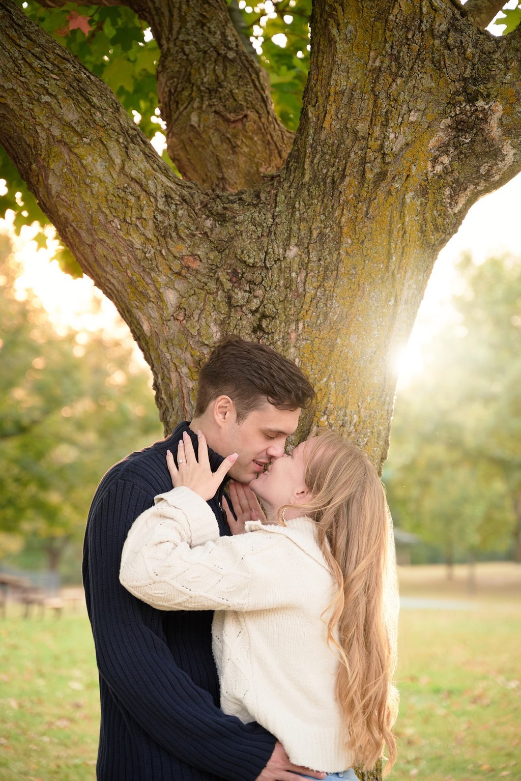 a man and woman kissing in front of a tree with sunset behind in a park