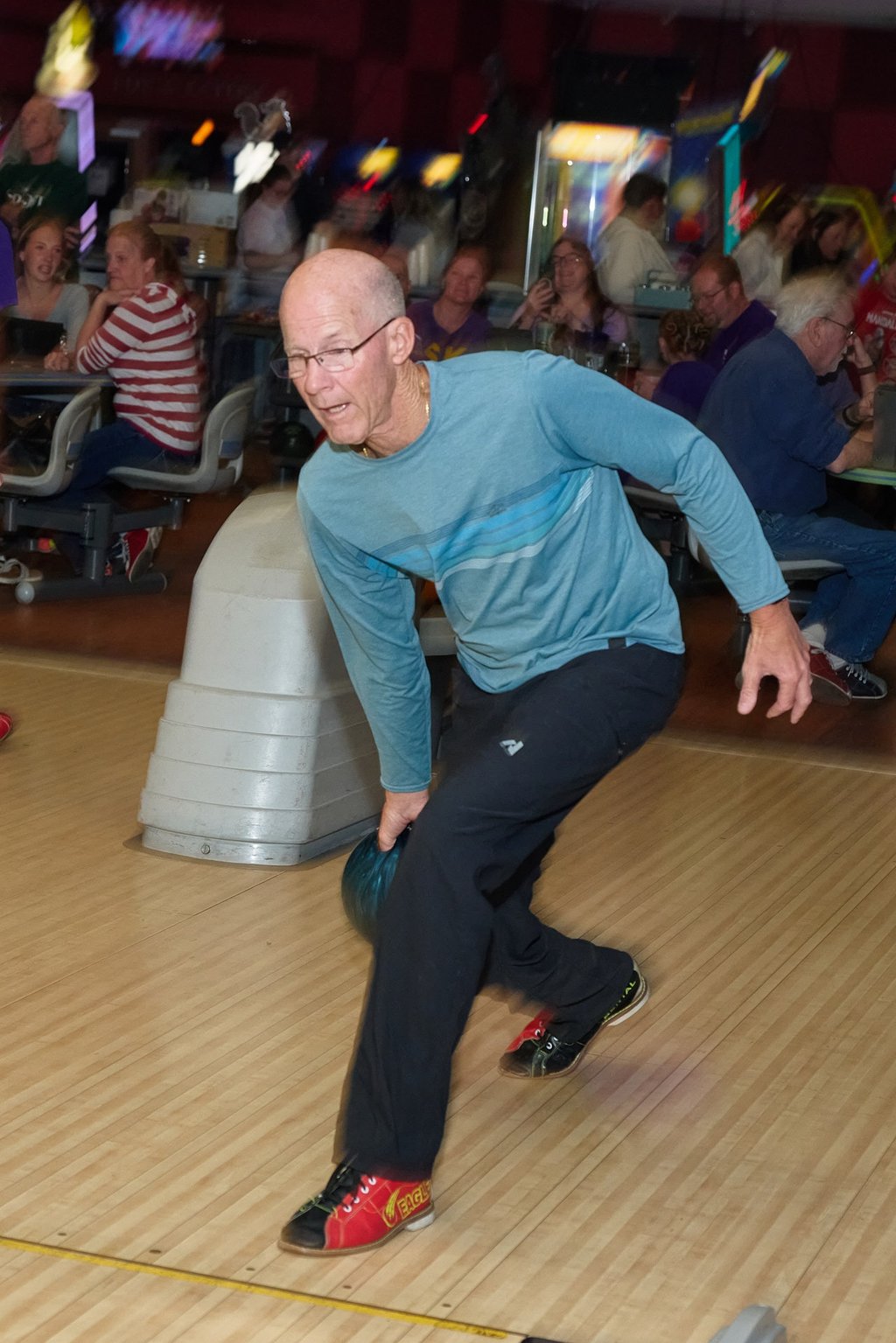 A senior man bowling in a teal shirt and black pants at a bowling alley with people in the background.
