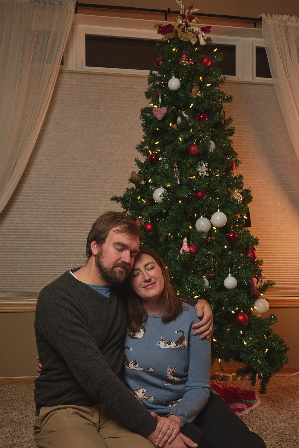 A couple hugging and sitting in front of a decorated Christmas tree with warm lights and ornaments.