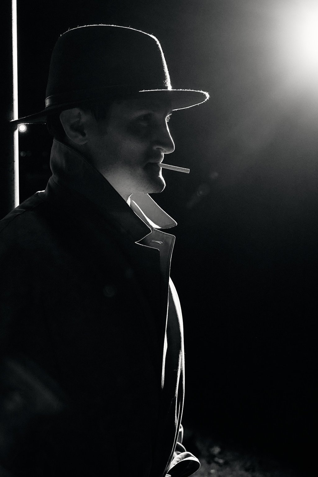 a man in a hat and a cigarette standing a dark area with street light from afar