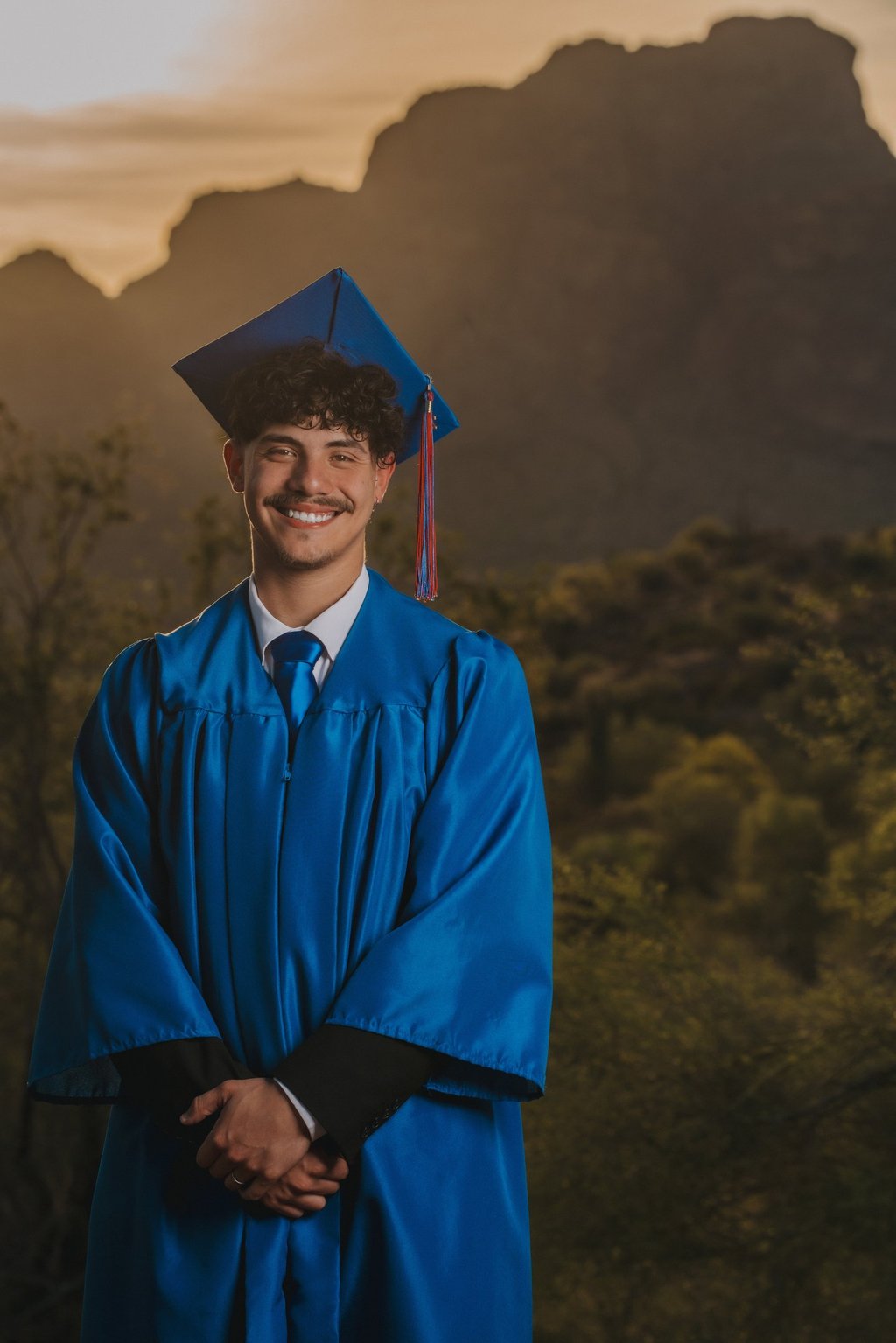 Smiling male graduate in blue cap and gown during a desert sunset graduation photoshoot.
