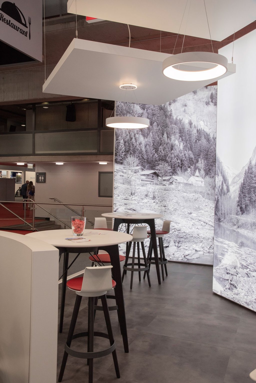 Lounge-Bereich mit Stehtischen und Hockern vor alpiner Bildwelt auf einer Messe.