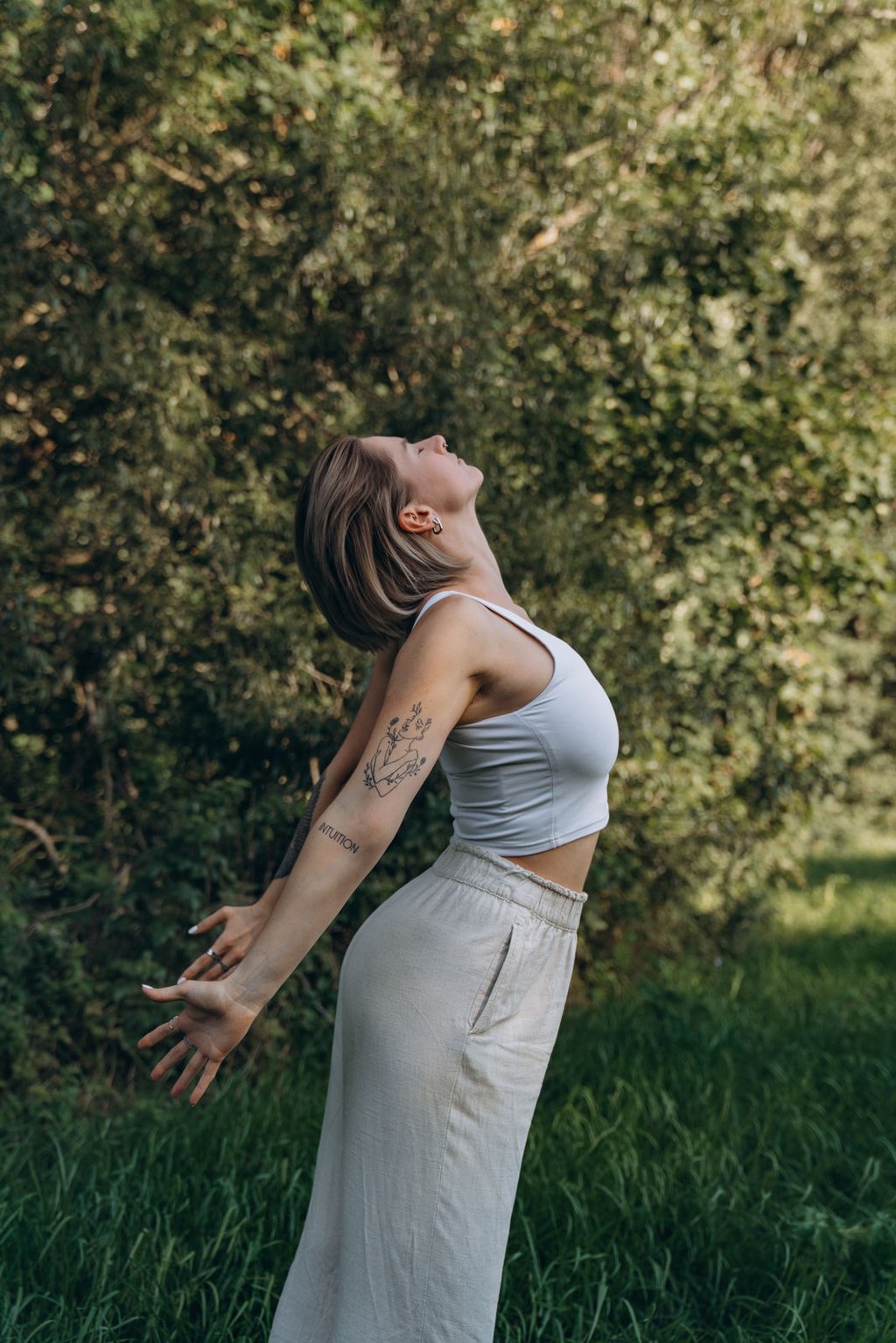 A young woman with tattoos practices mindful stretching in a lush green forest setting.