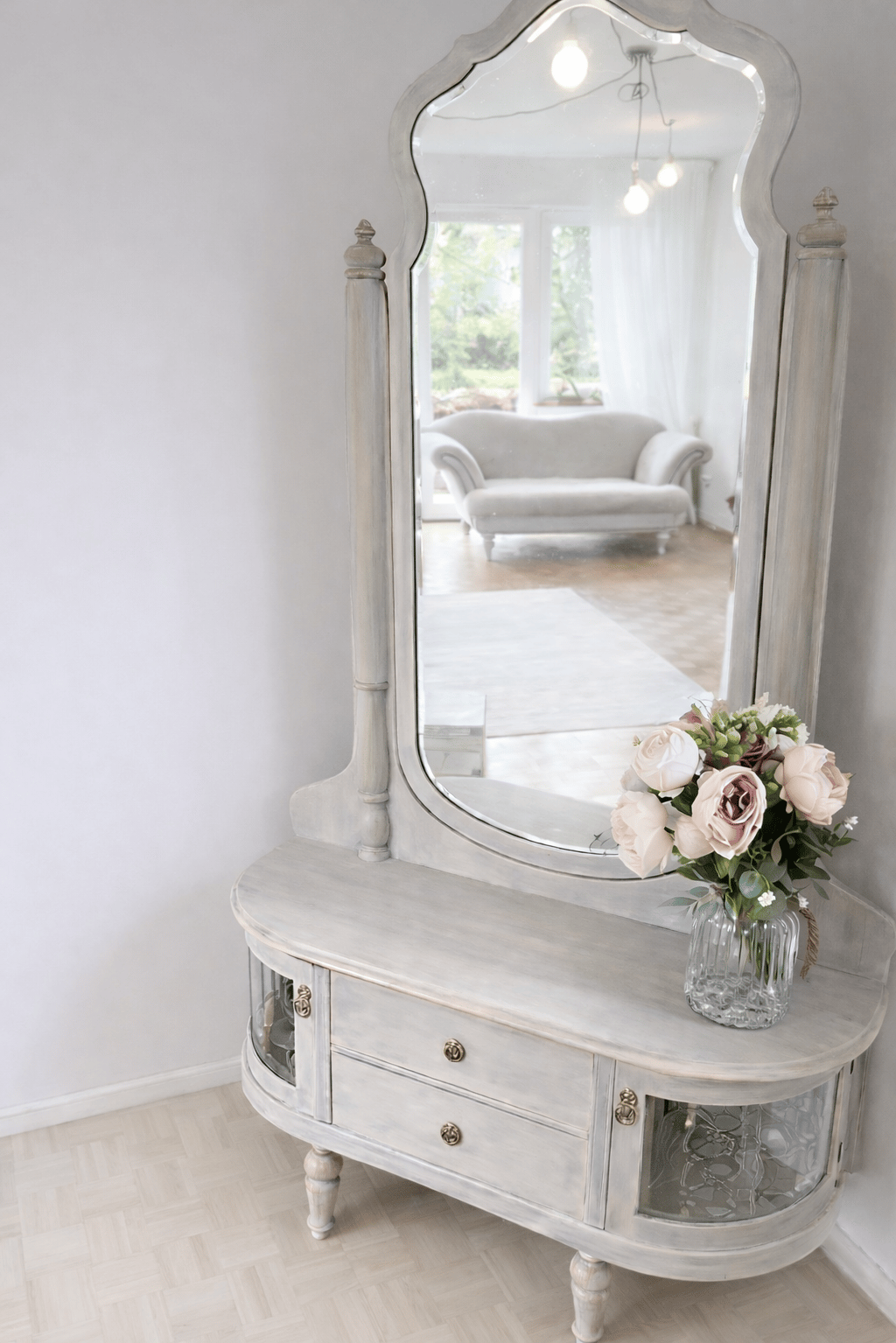 Vintage grey wooden vanity table with large mirror and bouquet of pink roses in a bedroom.