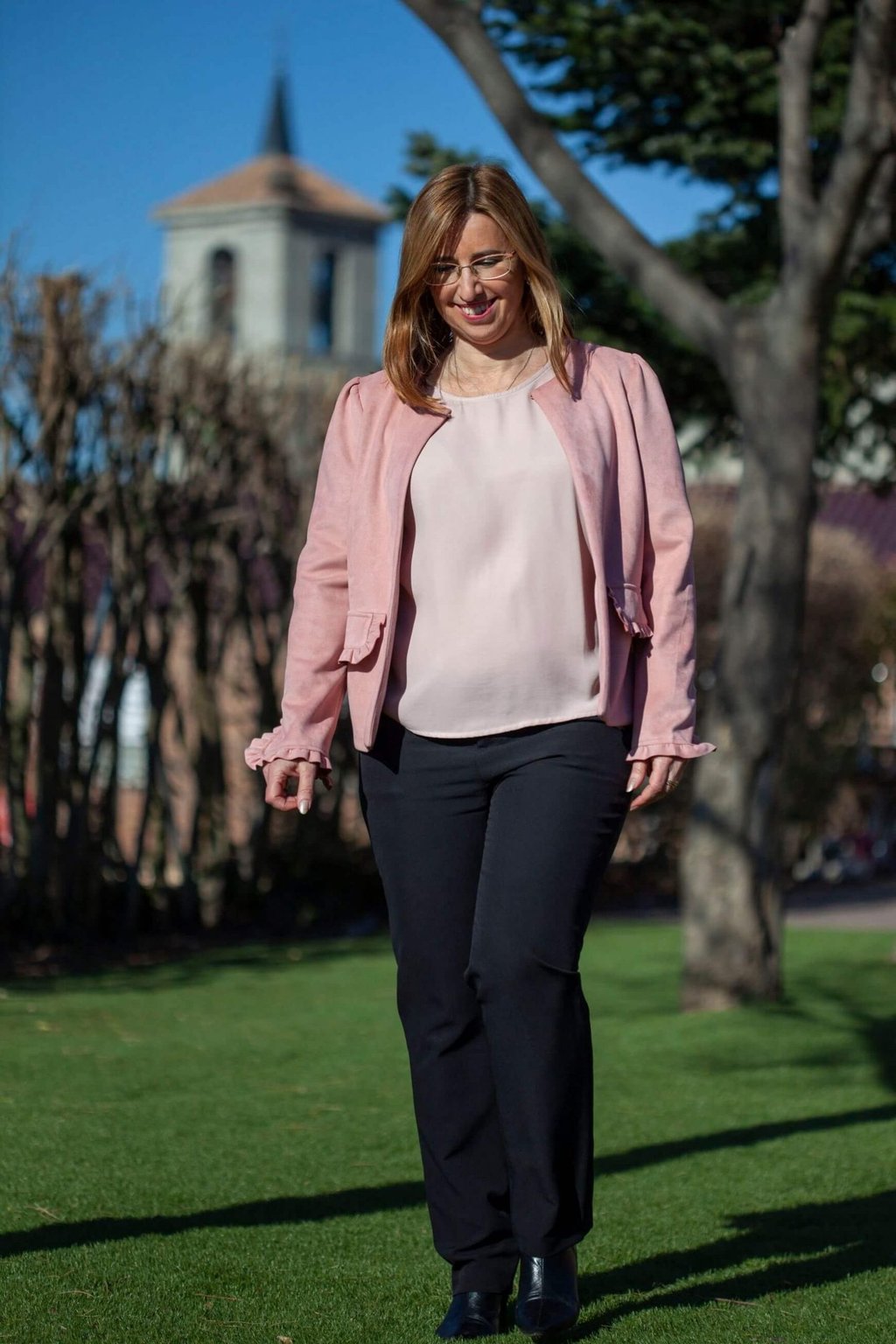 A woman wearing a stylish pink jacket and black trousers walking outdoors in a sunny park.