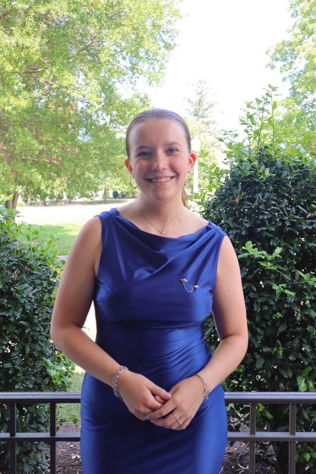 Smiling woman wearing a blue satin cowl neck dress standing outdoors in a garden setting.