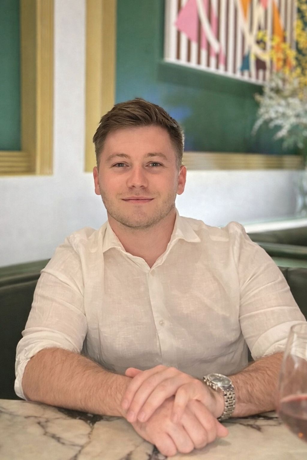 Harrison, smiling, wearing a white linen shirt and silver watch sitting at a restaurant table.