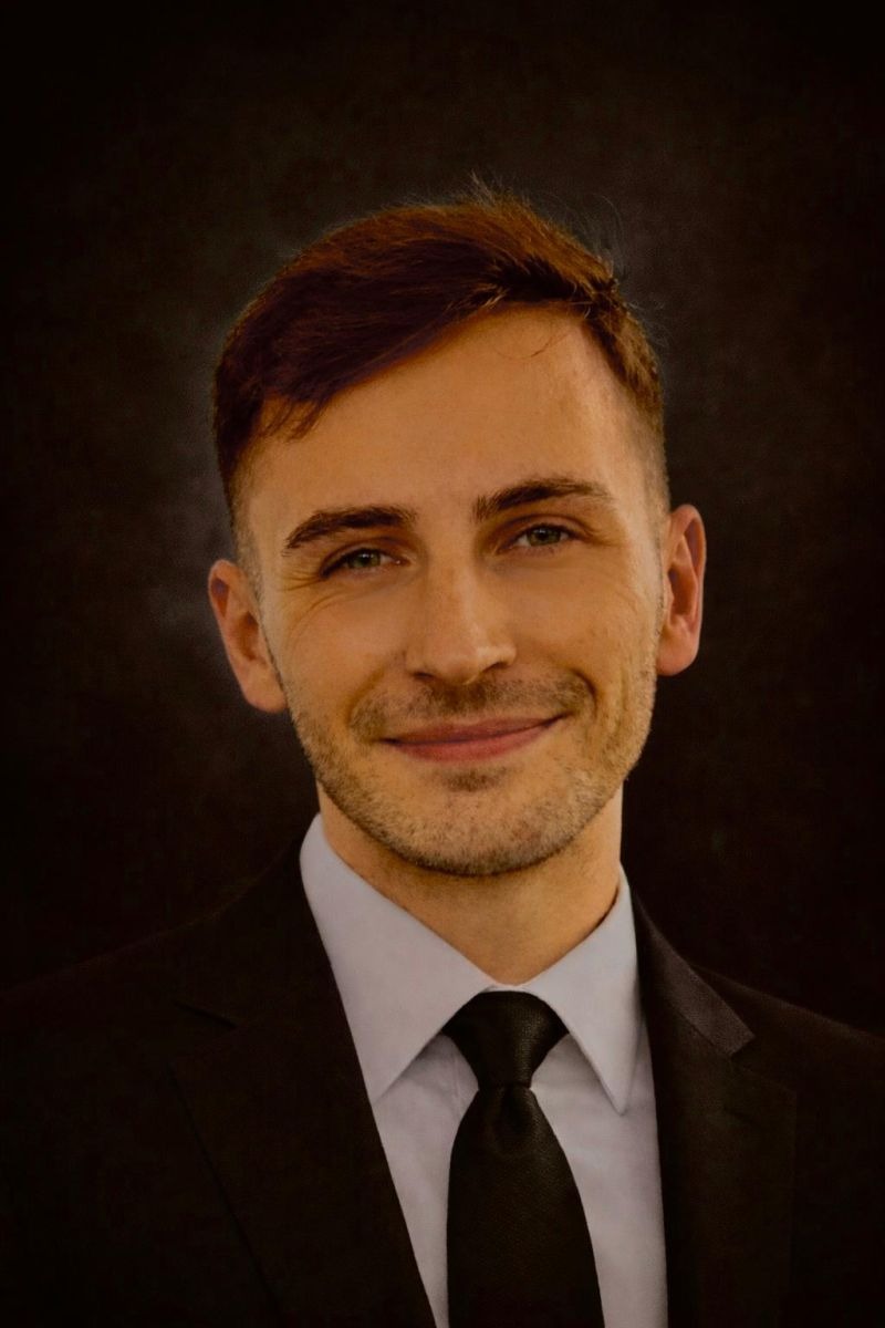 A smiling Bradley in a black suit and tie against a dark background.