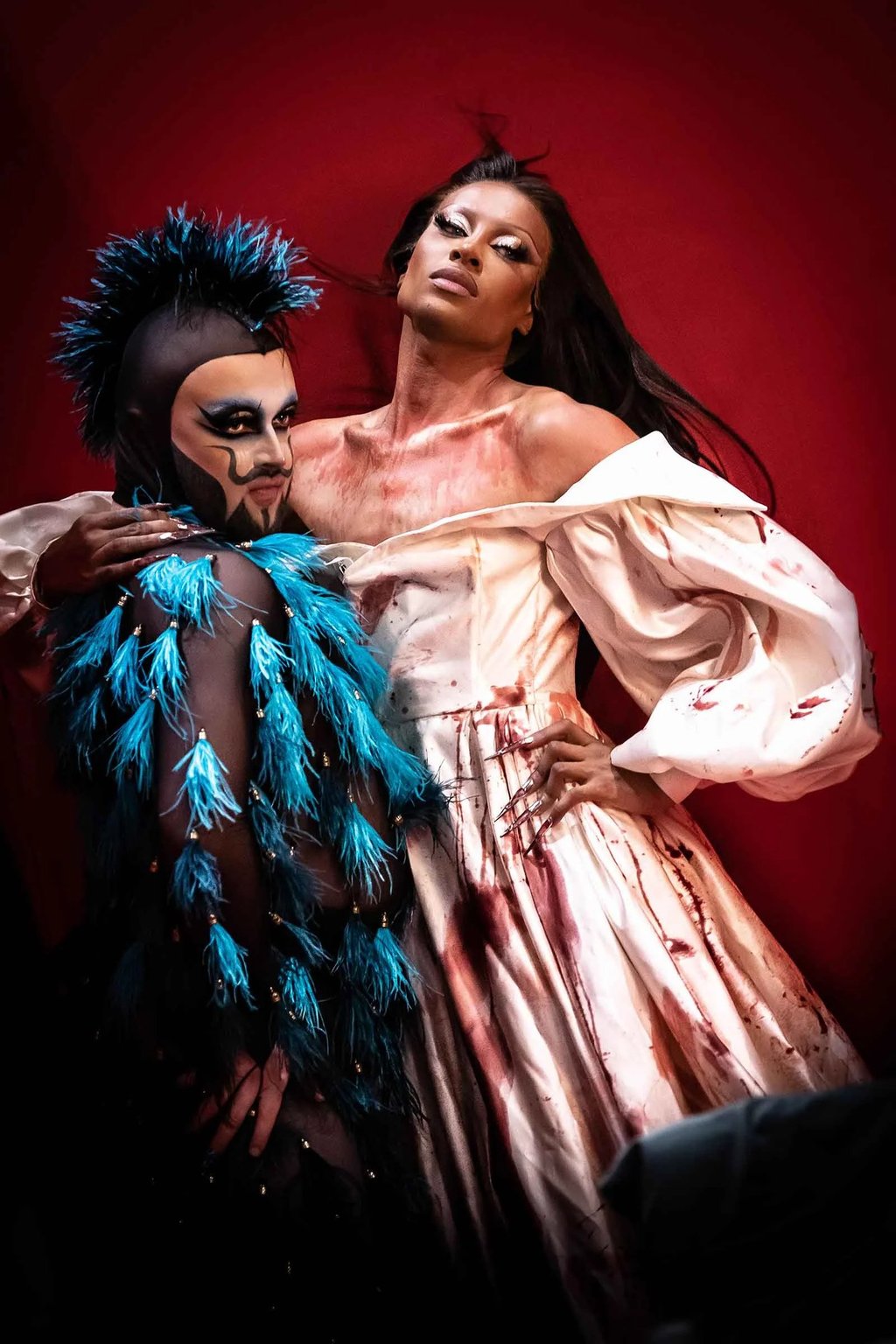 Two drag performers in artistic makeup and high-fashion horror costumes pose against a red background.