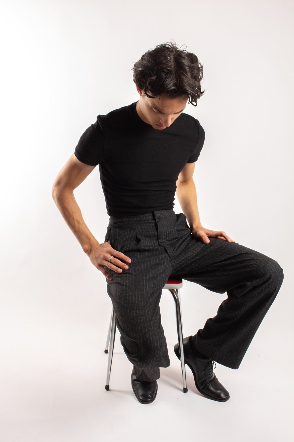 Young man in a black t-shirt and pinstripe trousers sitting on a stool for a fashion studio shoot.