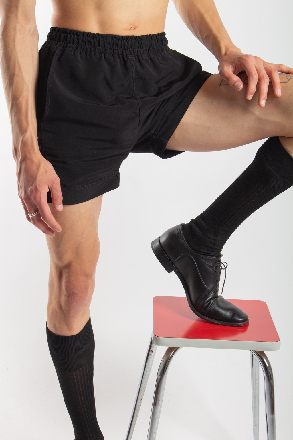 A man wearing black athletic shorts, dress shoes, and tall socks poses with one foot on a red stool.