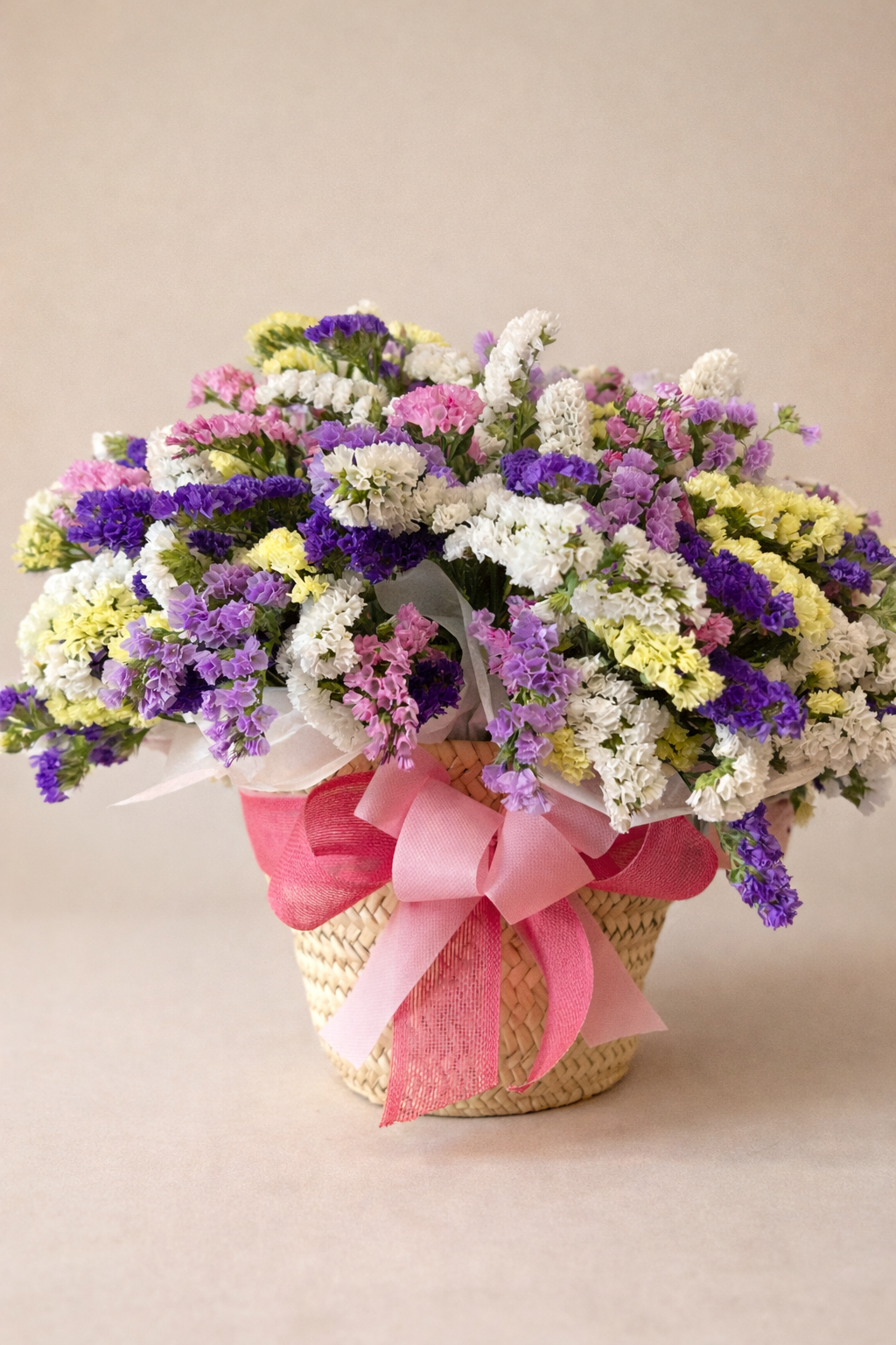A colorful bouquet of purple, pink, and white dried statice flowers in a woven basket with a pink ribbon.