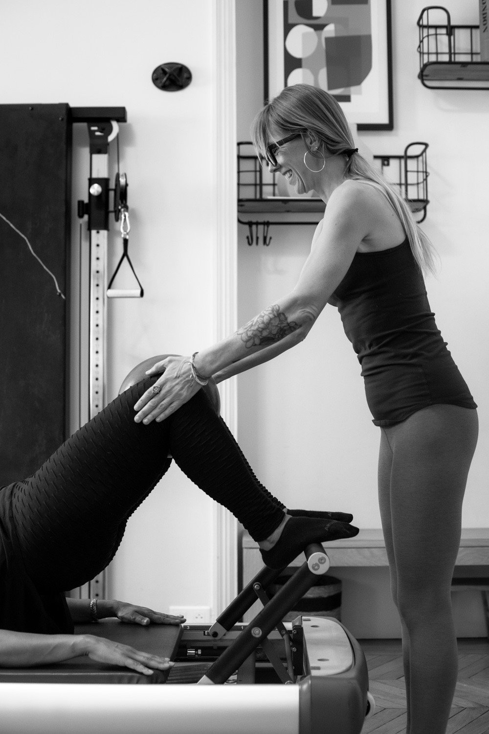 A smiling fitness instructor assists a client with a leg bridge exercise on a Pilates reformer machine.