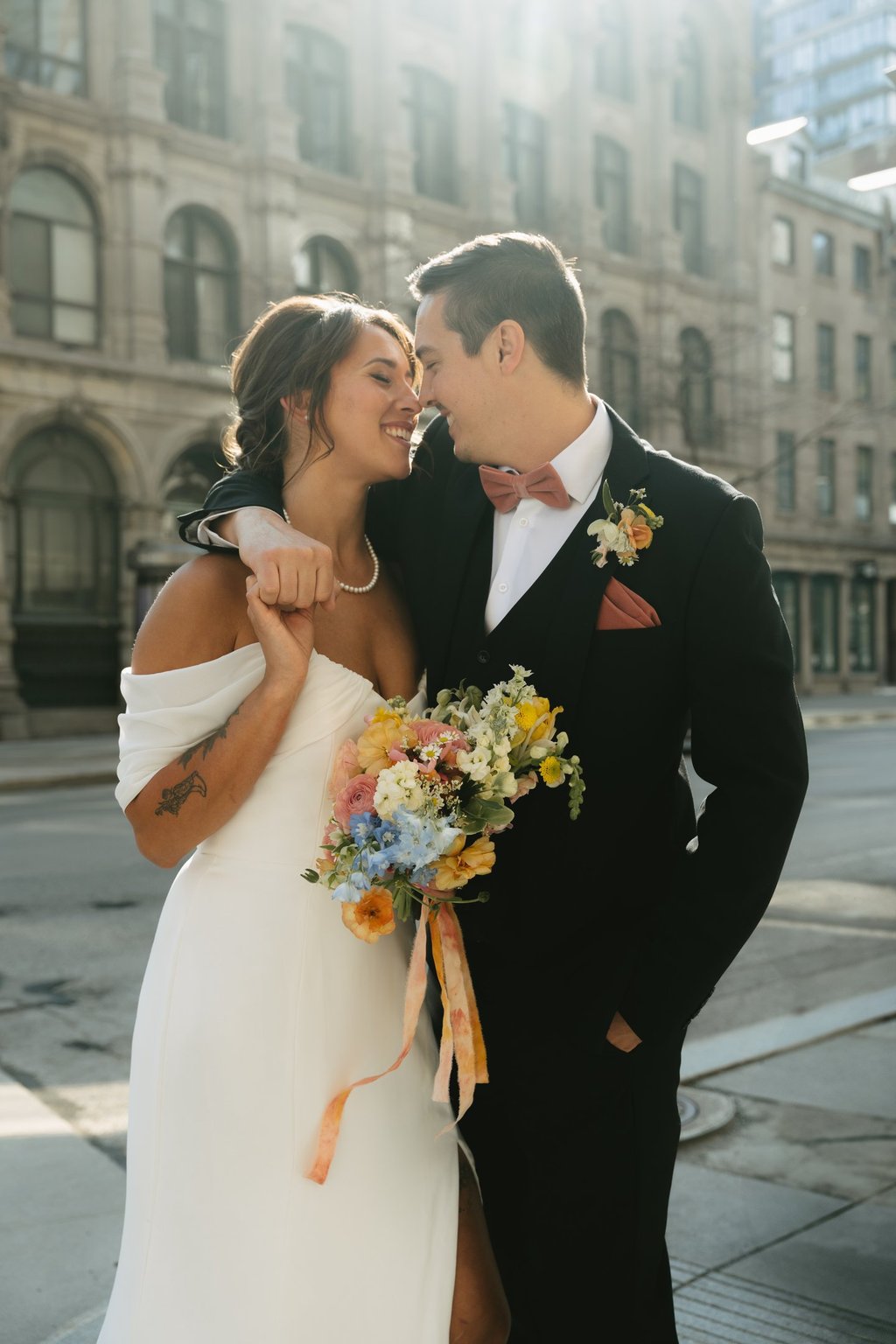 Une mariée et un marié souriants s’enlaçant dans une rue de la ville avec un bouquet coloré de fleur