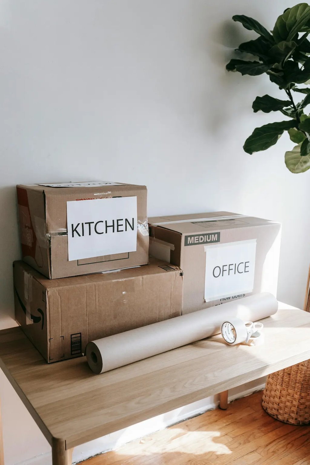 Labeled cardboard moving boxes for kitchen and office next to packing supplies on a wooden table.