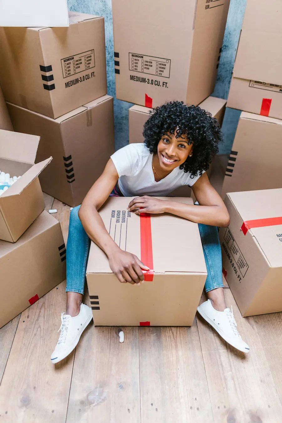 Smiling woman with curly hair sitting on the floor surrounded by cardboard moving boxes.