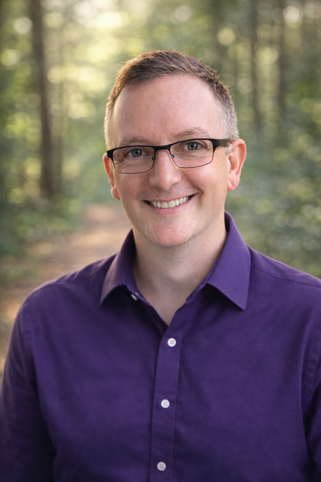 Smiling man with glasses in a purple shirt posing for a professional headshot in a forest.