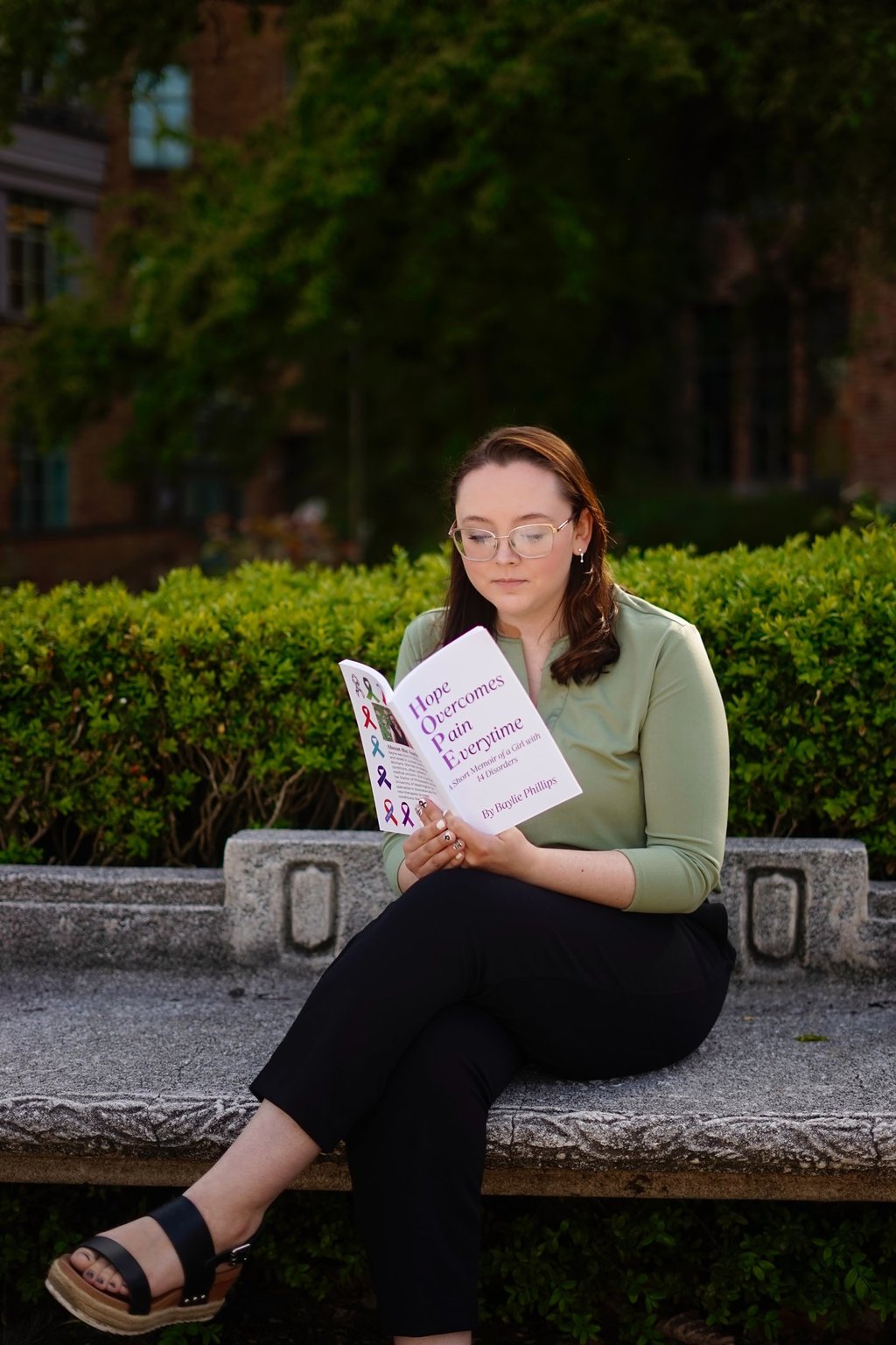Author Baylie Phillips sitting on a bench reading Hope Overcomes Pain Overtime book.