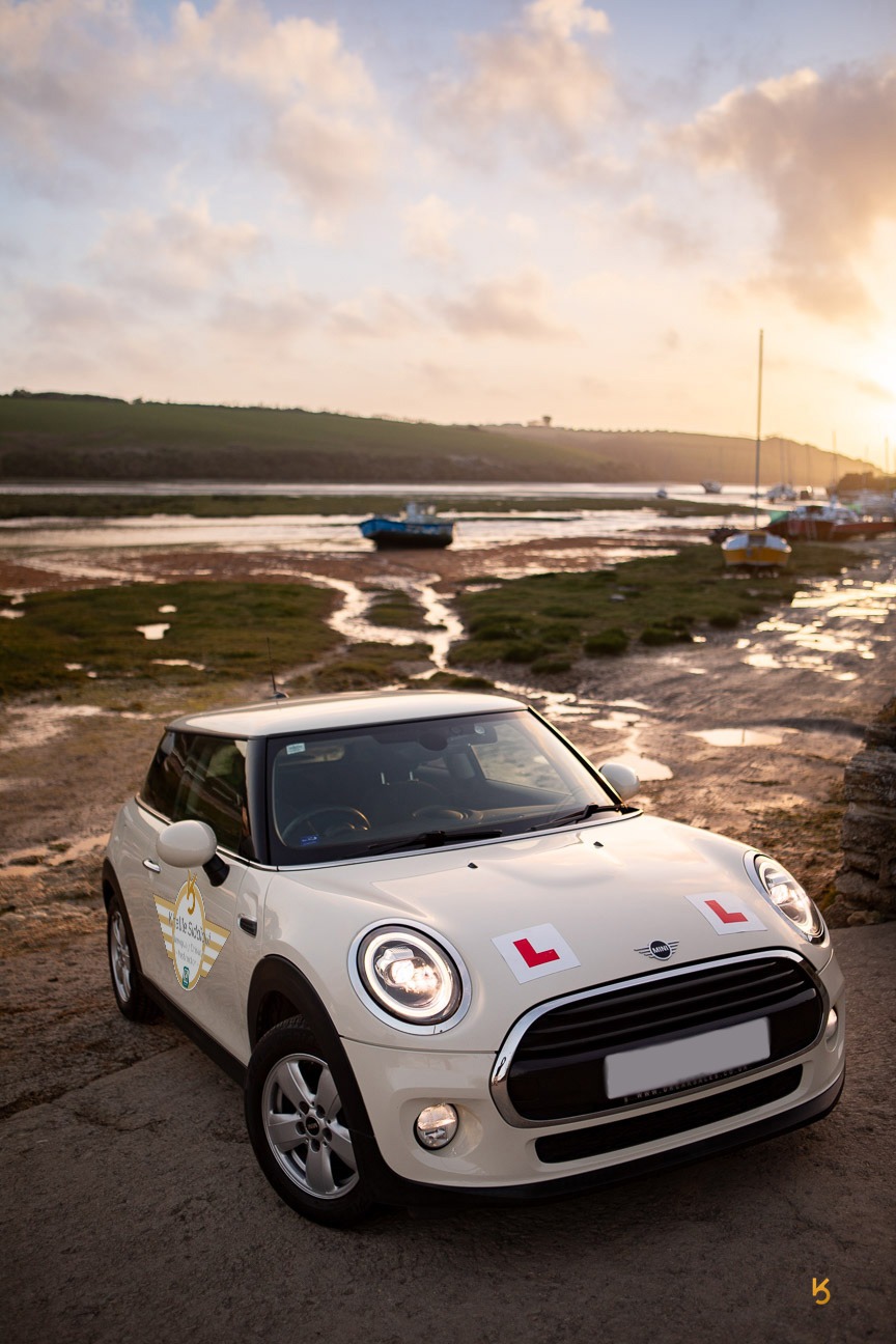 White Mini Cooper driving school car with L-plates parked at sunset by a scenic river estuary.